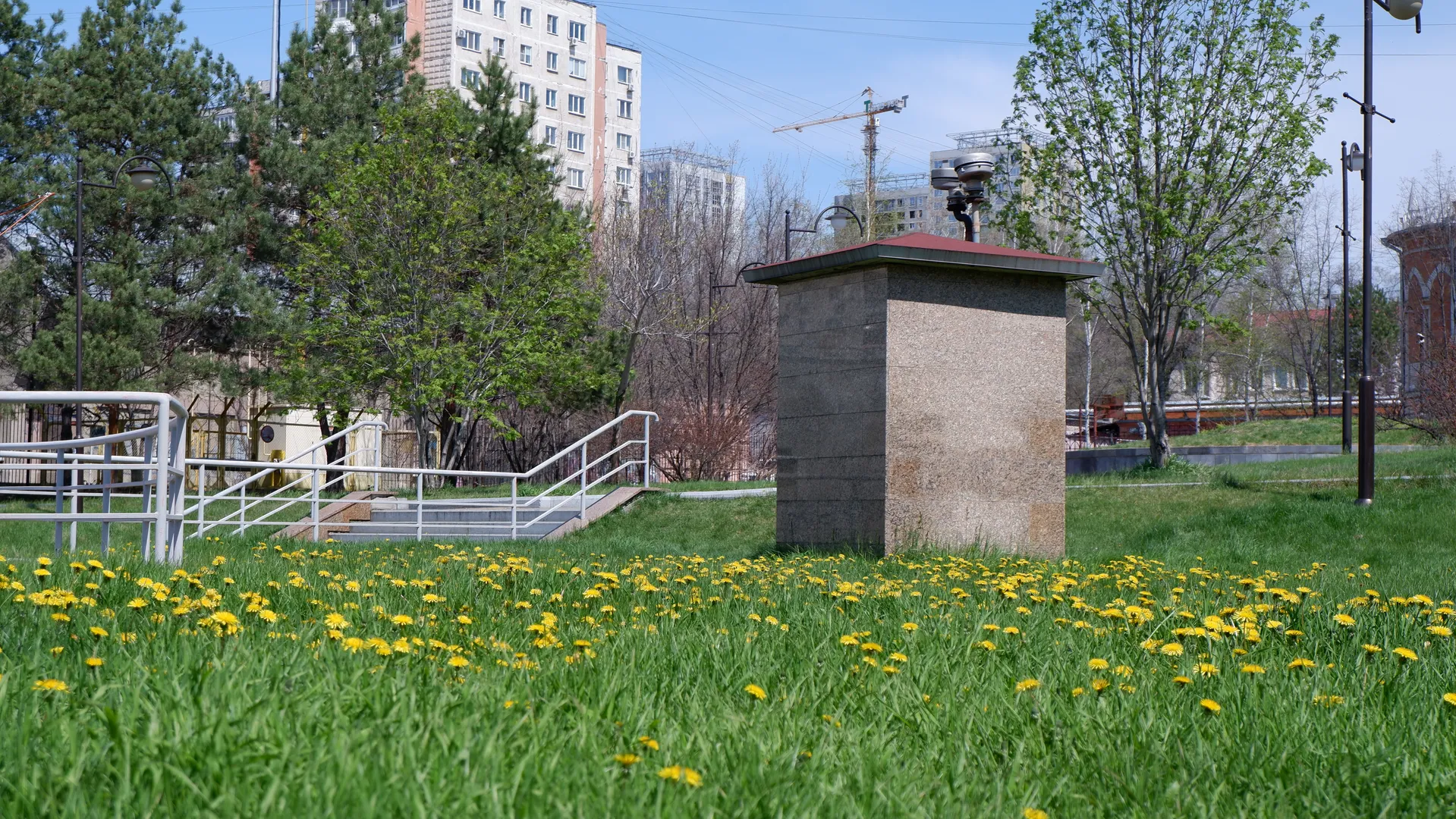 A stone structure stands on a grassy area, surrounded by trees and buildings. Yellow flowers are blooming on the grass, with a metal railing visible in the background. Cranes and multi-story buildings can be seen in the distance.