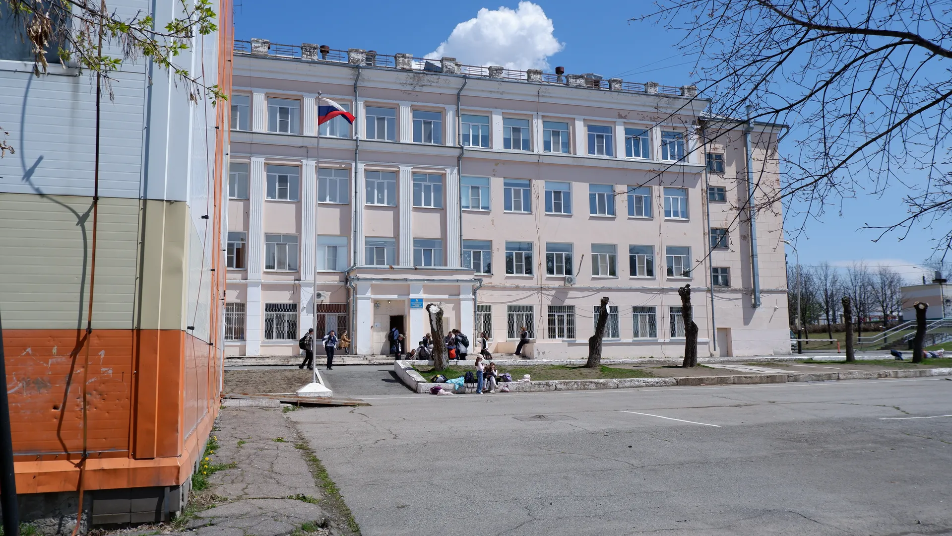 In front of a multi-story building is a square, with a flag flying atop the building. Several people are present in front of the building, and a leafless tree stands nearby. The sky is clear and blue.