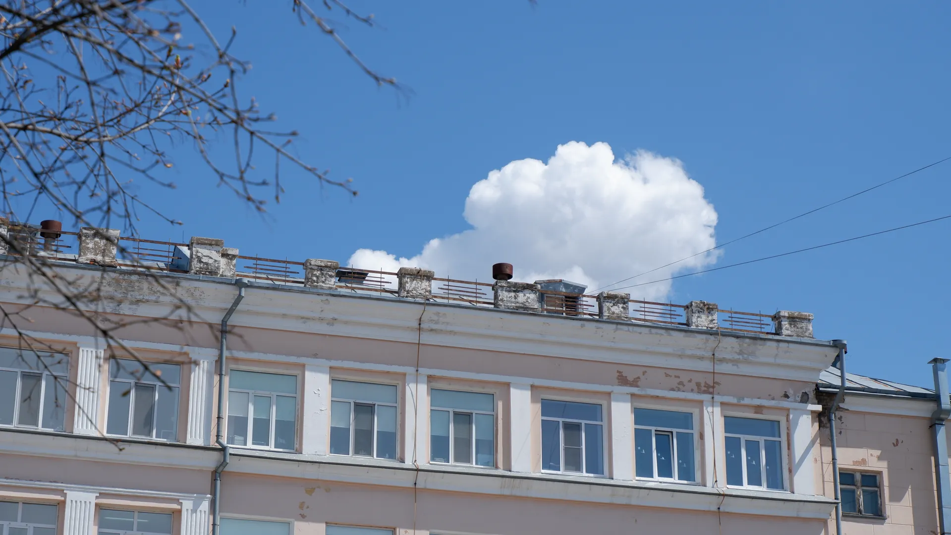 The top of a multi-story building has several ventilation pipes and railings. The facade has multiple light-colored windows. A few white clouds are in the sky, with branches partially blocking the view.