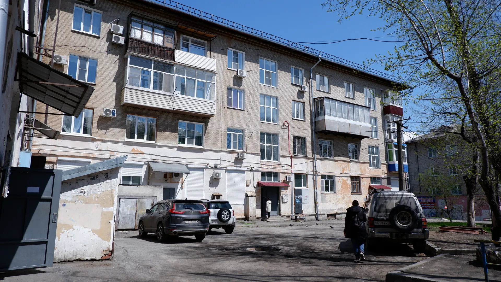 Several cars are parked in front of a multi-story residential building, which has balconies and air conditioning units. A person is walking nearby, with trees providing shade. The building looks somewhat aged, with peeling on the walls.