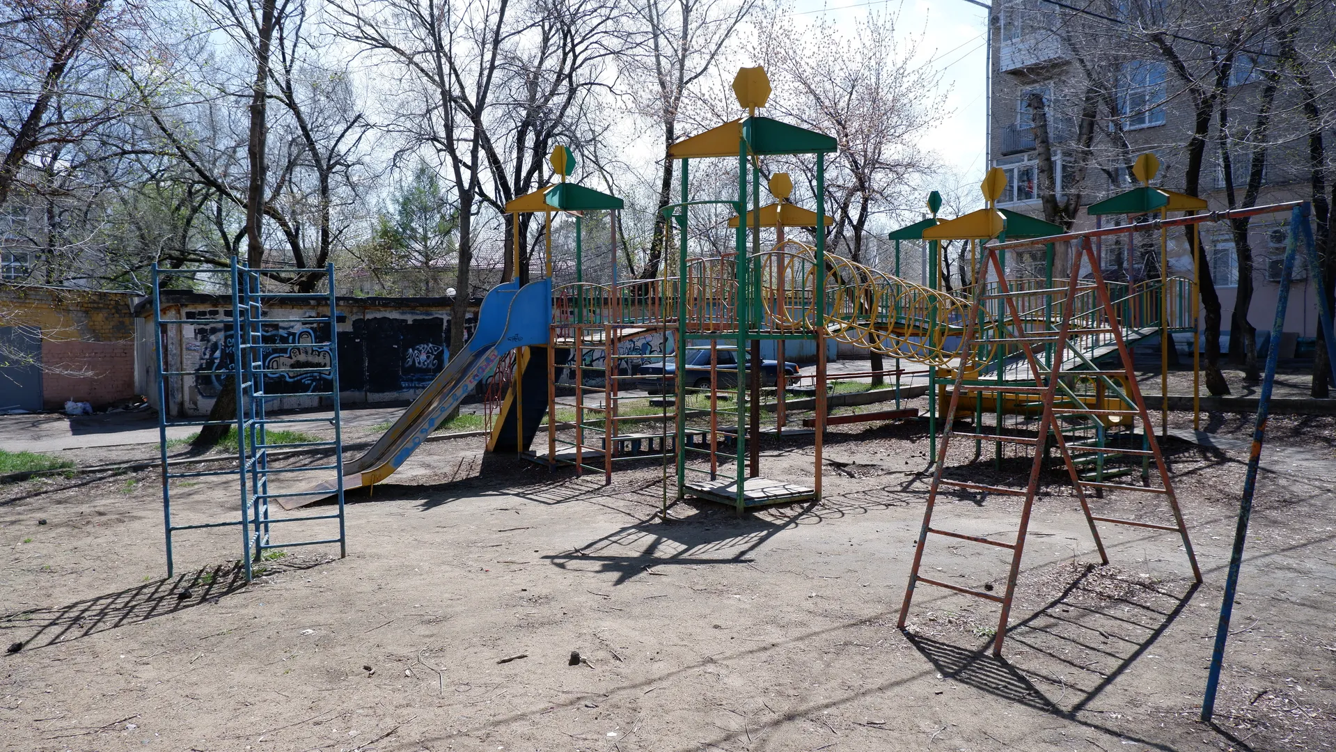 A children’s playground features slides, climbing frames, and swings. The ground is covered with sand, and trees surround the area. Buildings and parked vehicles are visible in the background.