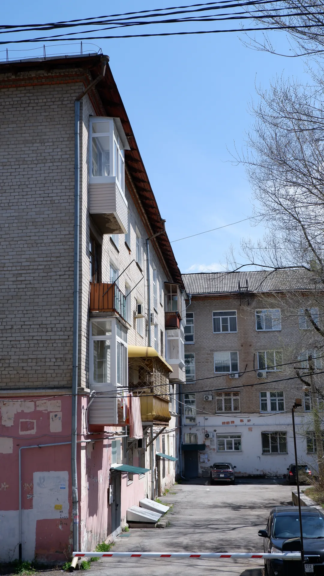 A narrow road lies between two multi-story buildings, with a parking barrier on the road. The facades have balconies and windows, with light-colored exteriors. The sky is clear, and power lines cross the scene.