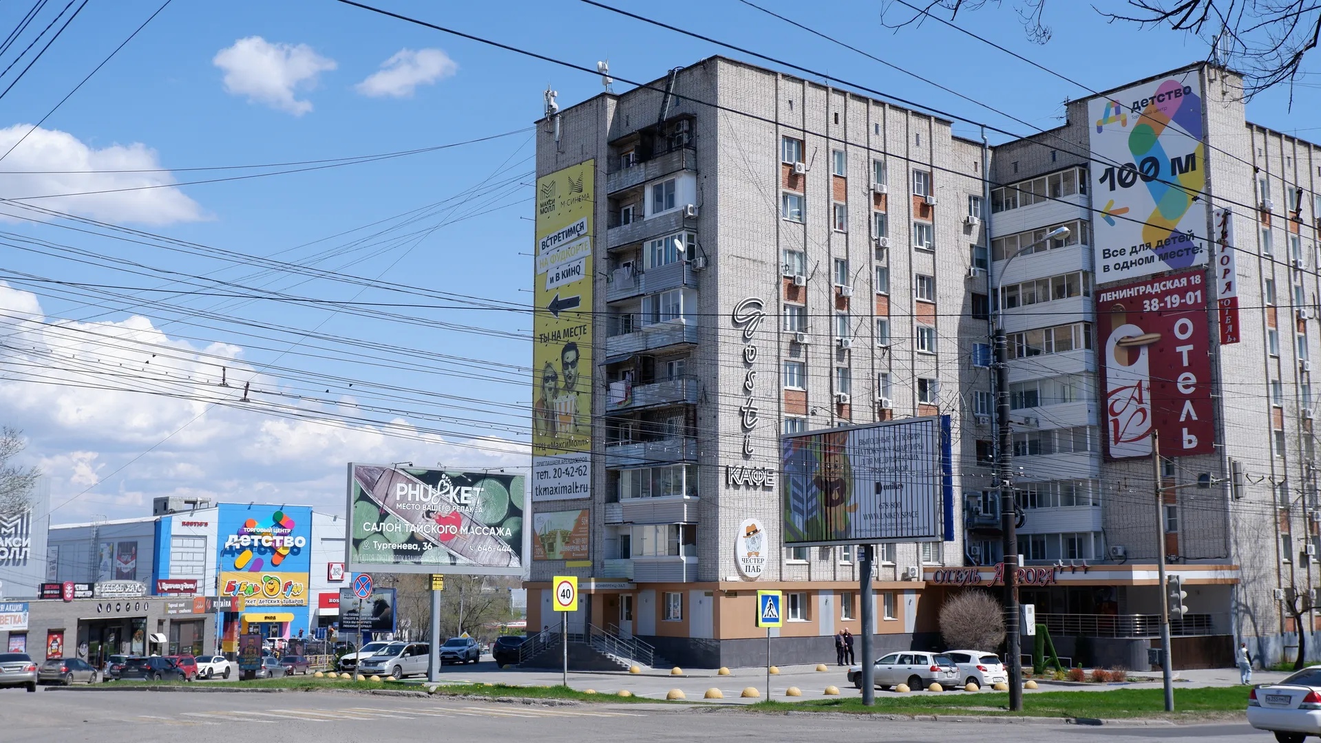 A street runs alongside multi-story buildings, which feature billboards and signs. Vehicles are driving on the street, along with traffic signs. A few white clouds dot the sky, with power lines crisscrossing overhead.