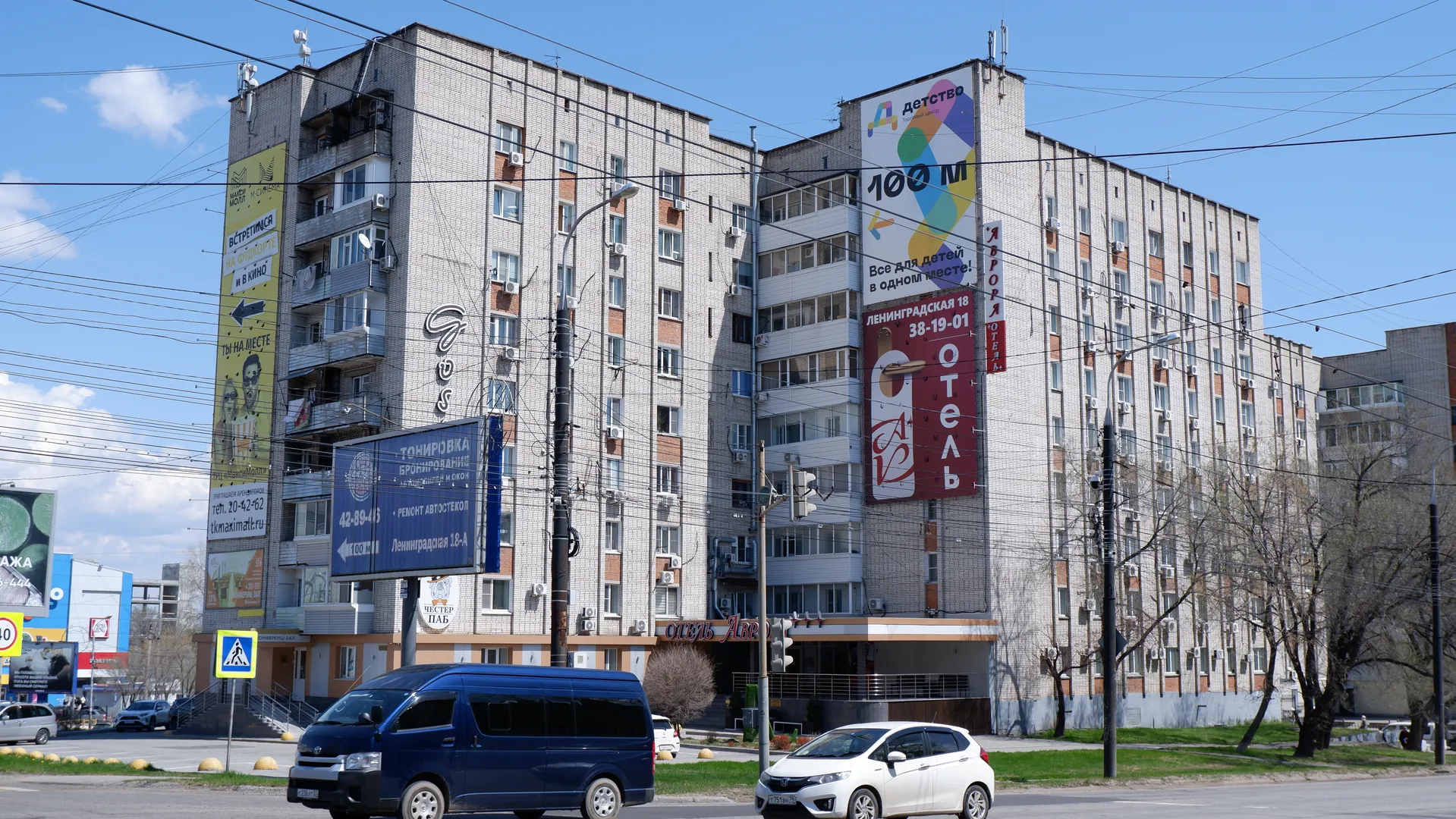 Vehicles travel along a street in front of multi-story buildings that display large billboards. The billboards contain commercial information in bold fonts. The sky is clear, with power lines spanning the scene.