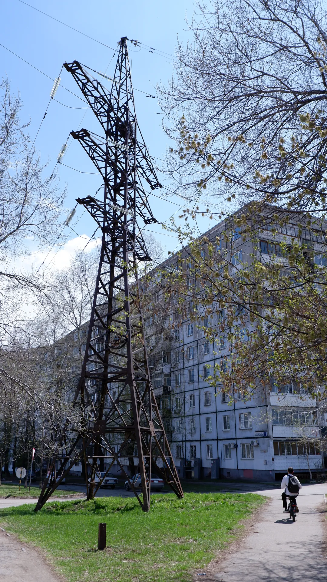 A tall metal power tower stands in the foreground, with a multi-story building and trees in the background. The building has neatly arranged windows and a light-colored exterior. A person is riding a bicycle past the right side of the tower.