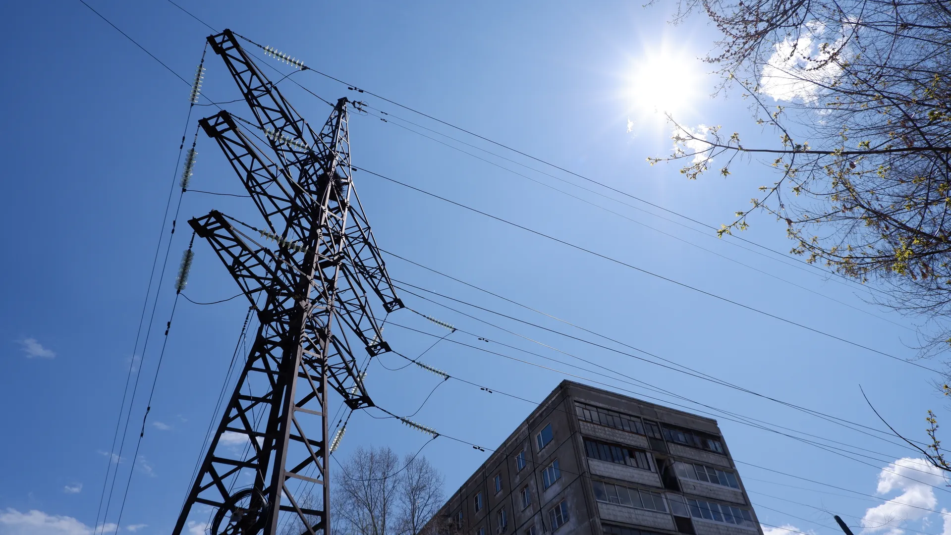 The metal power tower has a complex structure, with multiple power lines extending from it. The sky is clear and the sun is bright, illuminating the tower and nearby buildings. The windows of the buildings reflect the sunlight.