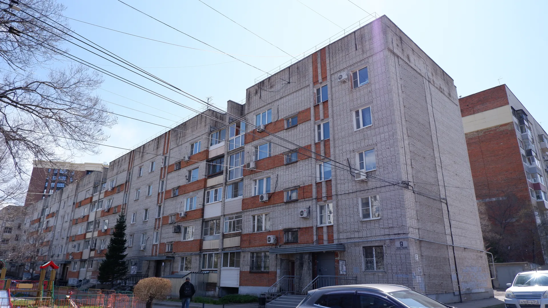 A multi-story residential building with a brick exterior, with some red-colored sections. Several cars are parked in front of the building, with trees and shrubs nearby. A person stands on the open area in front of the building.