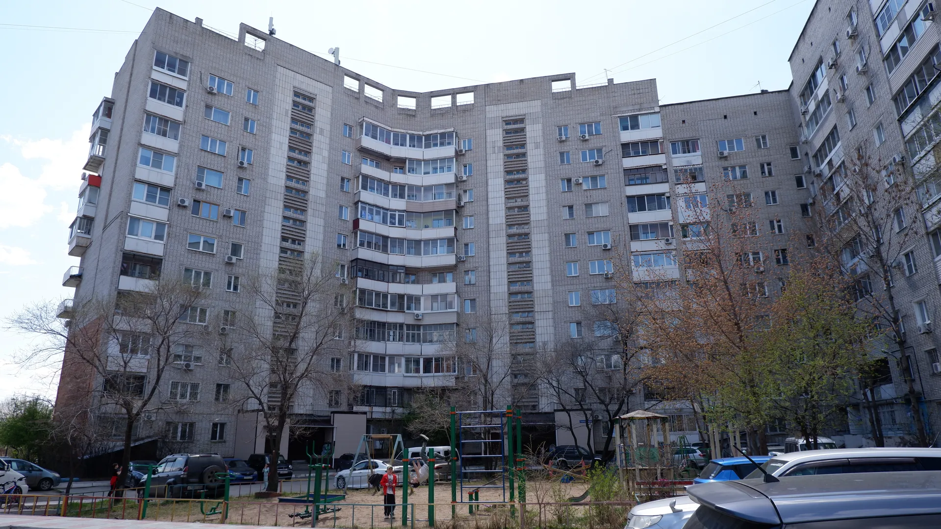 A high-rise building with a gray tiled exterior, featuring many windows and balconies. In front of the building is a children’s playground with slides and swings. Trees surround the building.