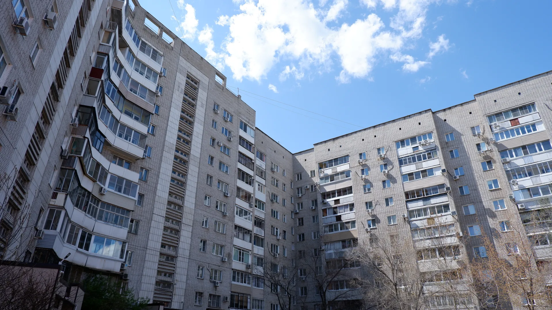 Two high-rise buildings form a U-shaped structure, with gray tiled facades and many windows. Some sparse trees grow between the buildings. A few white clouds are in the sky.
