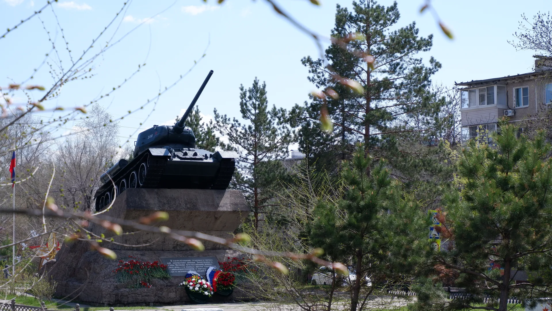 A tank memorial stands in a flower bed, with the tank’s barrel pointed towards the sky. Several tall pine trees with dense foliage surround the memorial. In the background, there is a building with bright windows.