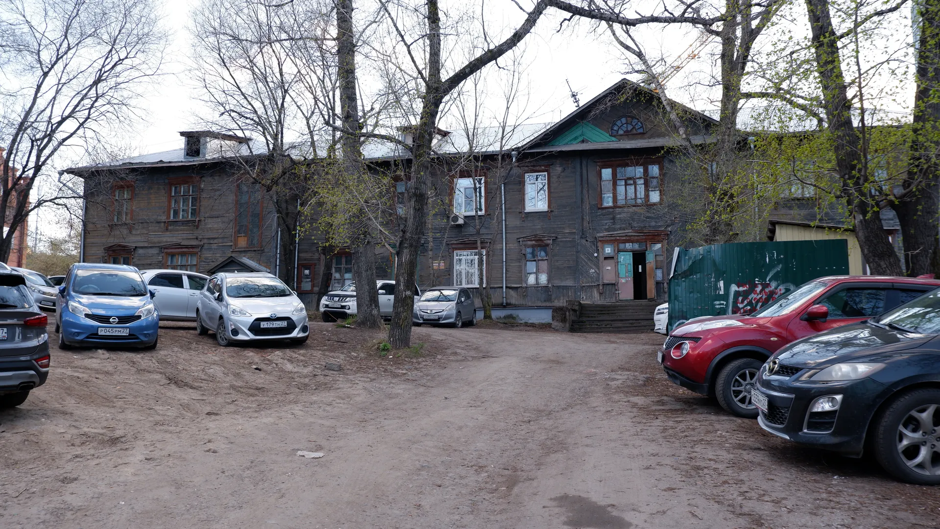 An old wooden building with a dark exterior and numerous windows. Several cars are parked in front of the building, with the ground made of dirt. A few trees with bare branches surround the building.