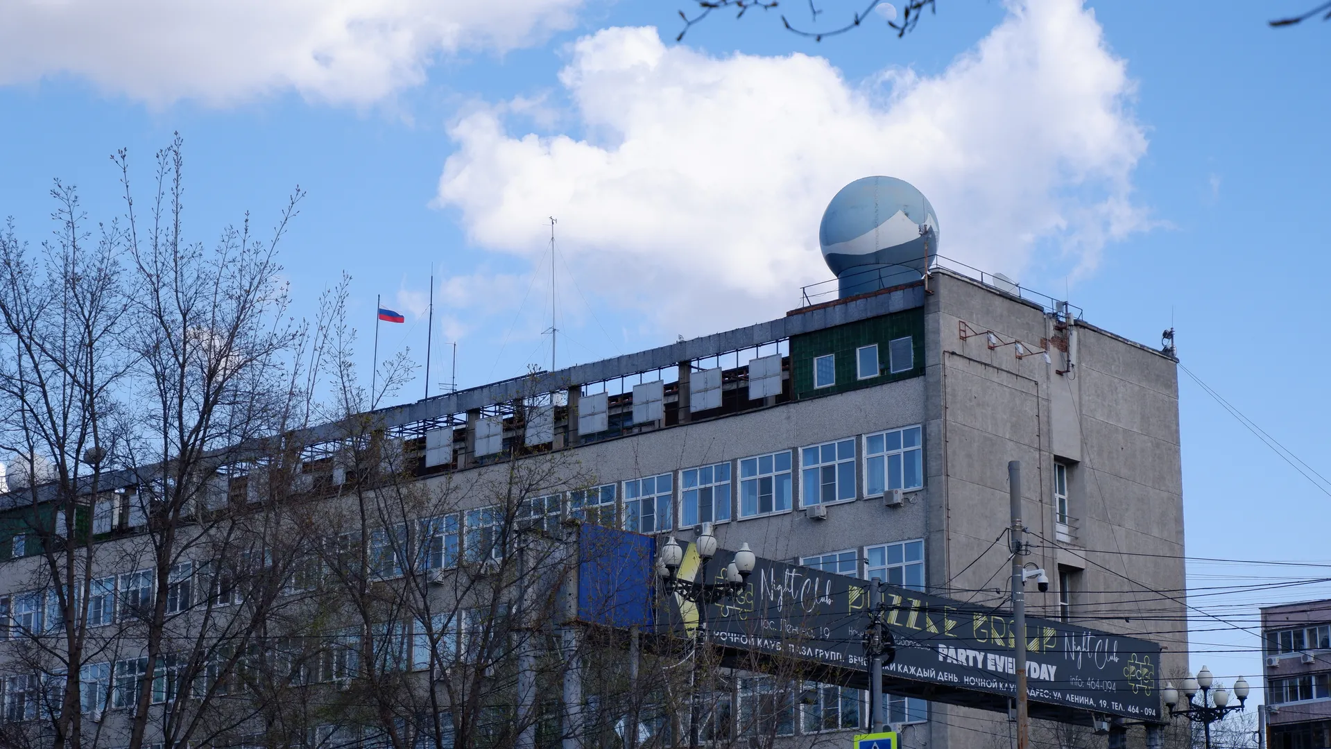 A high-rise building with a spherical structure on the top and a gray tiled exterior. The building’s front has multiple windows reflecting light. In front of the building, there are a few trees with bare branches, and white clouds can be seen in the sky.