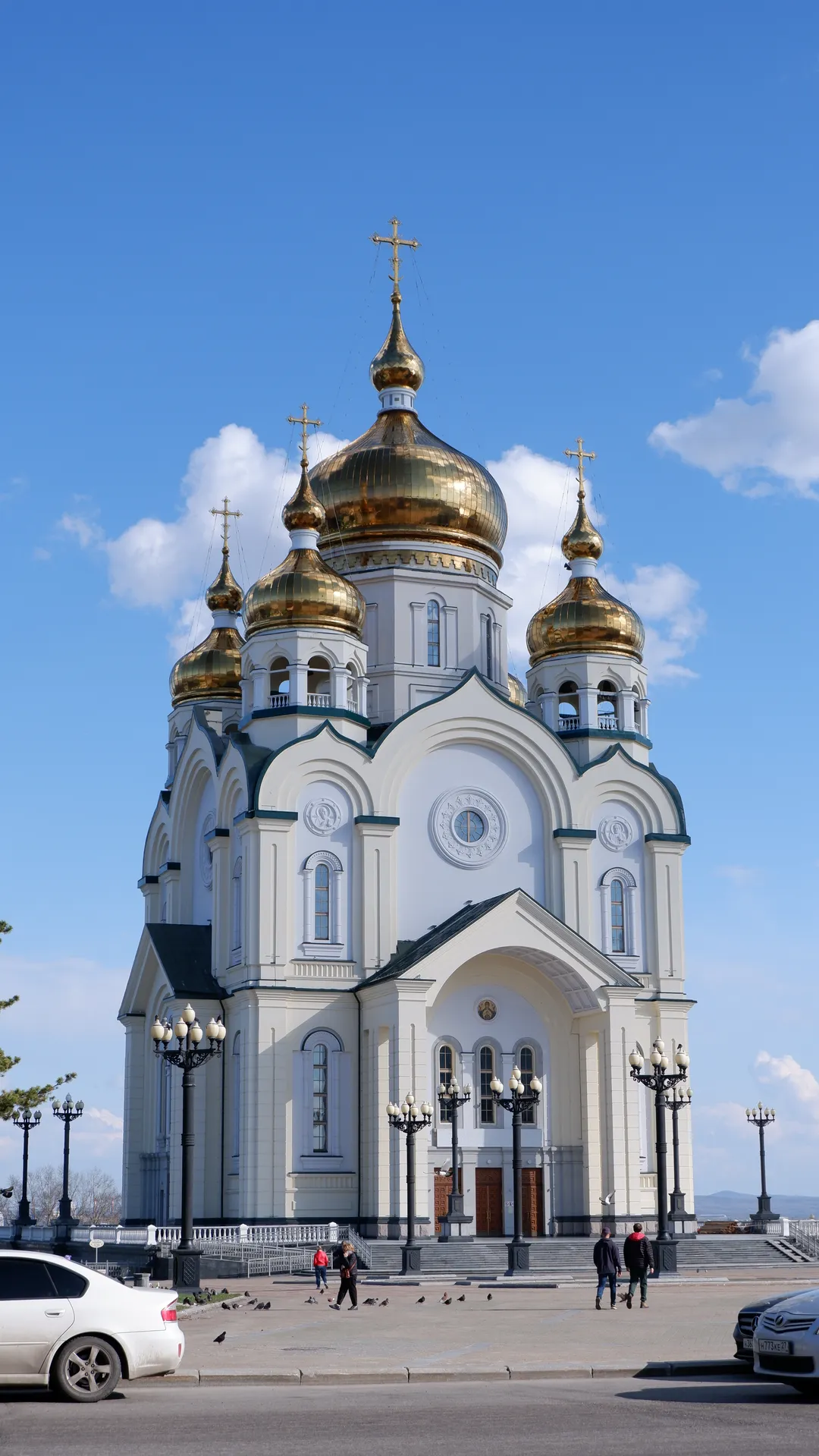 A white church with five golden domes and crosses on top. The church’s front features arched windows and decorative round patterns. Pedestrians and street lamps can be seen in the square in front of the church.