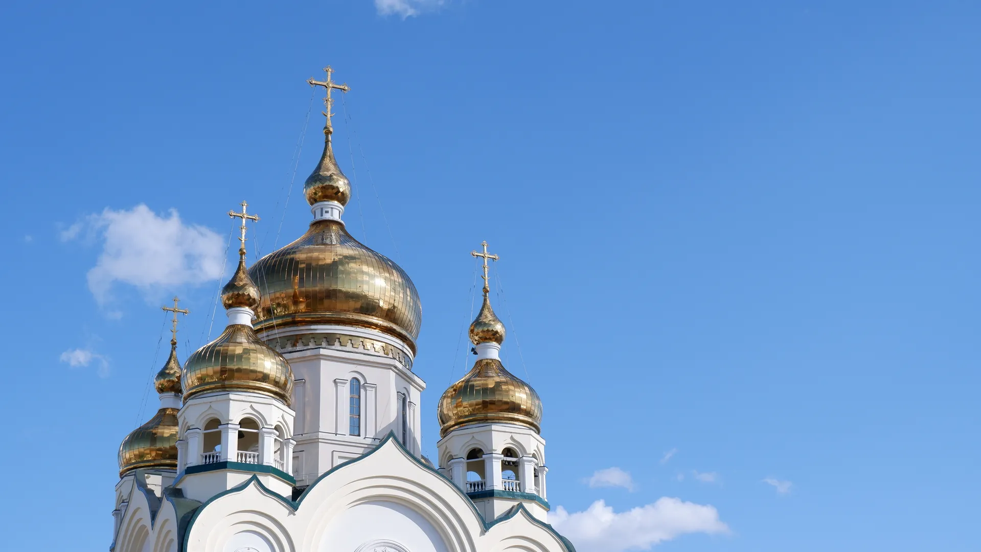 The church’s top has five golden domes and crosses, set against a backdrop of blue skies and white clouds. The church building is white with green decorative lines. There is ample empty space on the right side of the image.