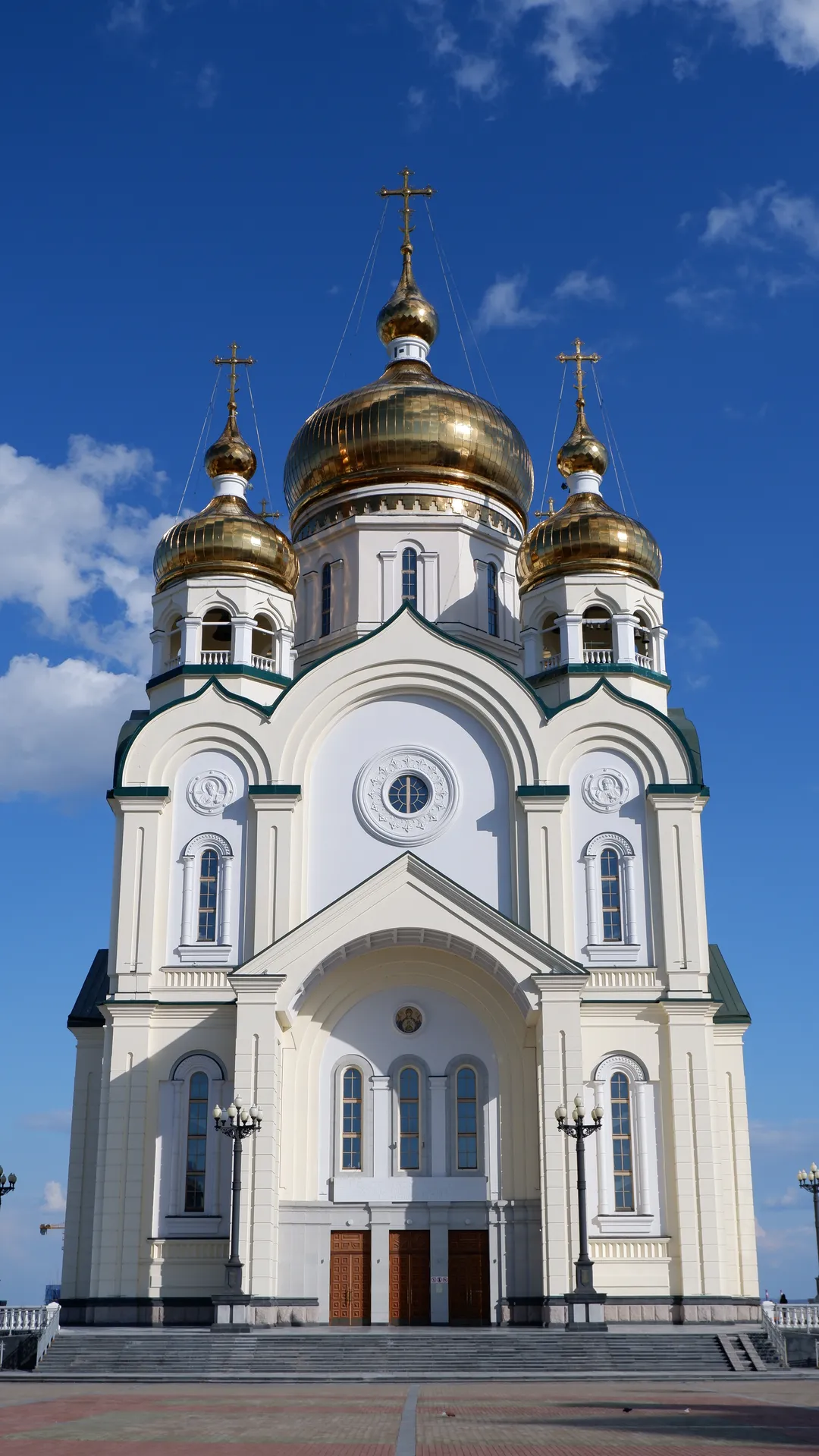 A white church with three golden domes and crosses on top. The church’s front has multiple arched windows and three large doors. Stairs and street lamps are visible in front of the church.