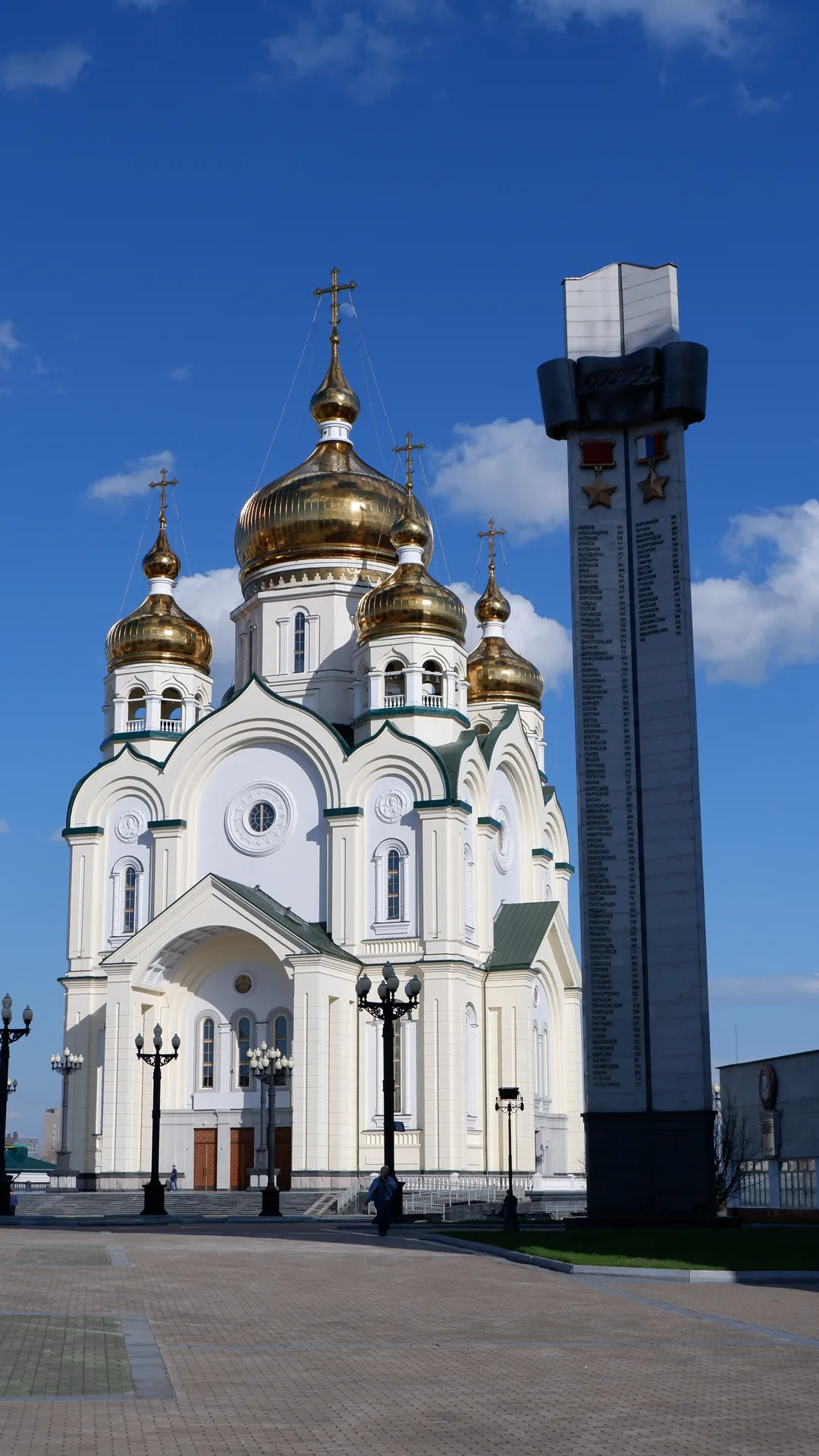 A white church with four golden domes and crosses on top. A tall monument with engraved text and stars stands beside the church. Pedestrians and street lamps are seen in the square in front of the church.