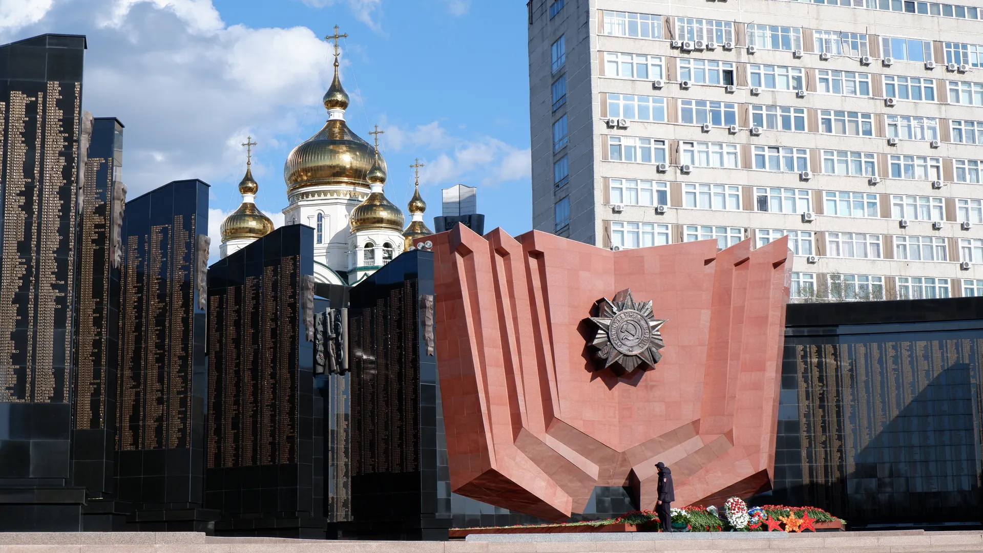 A red monument with a unique shape and a badge on top. On both sides of the monument are black stone slabs with engraved text. In the background, a golden-domed church and high-rise buildings are visible.
