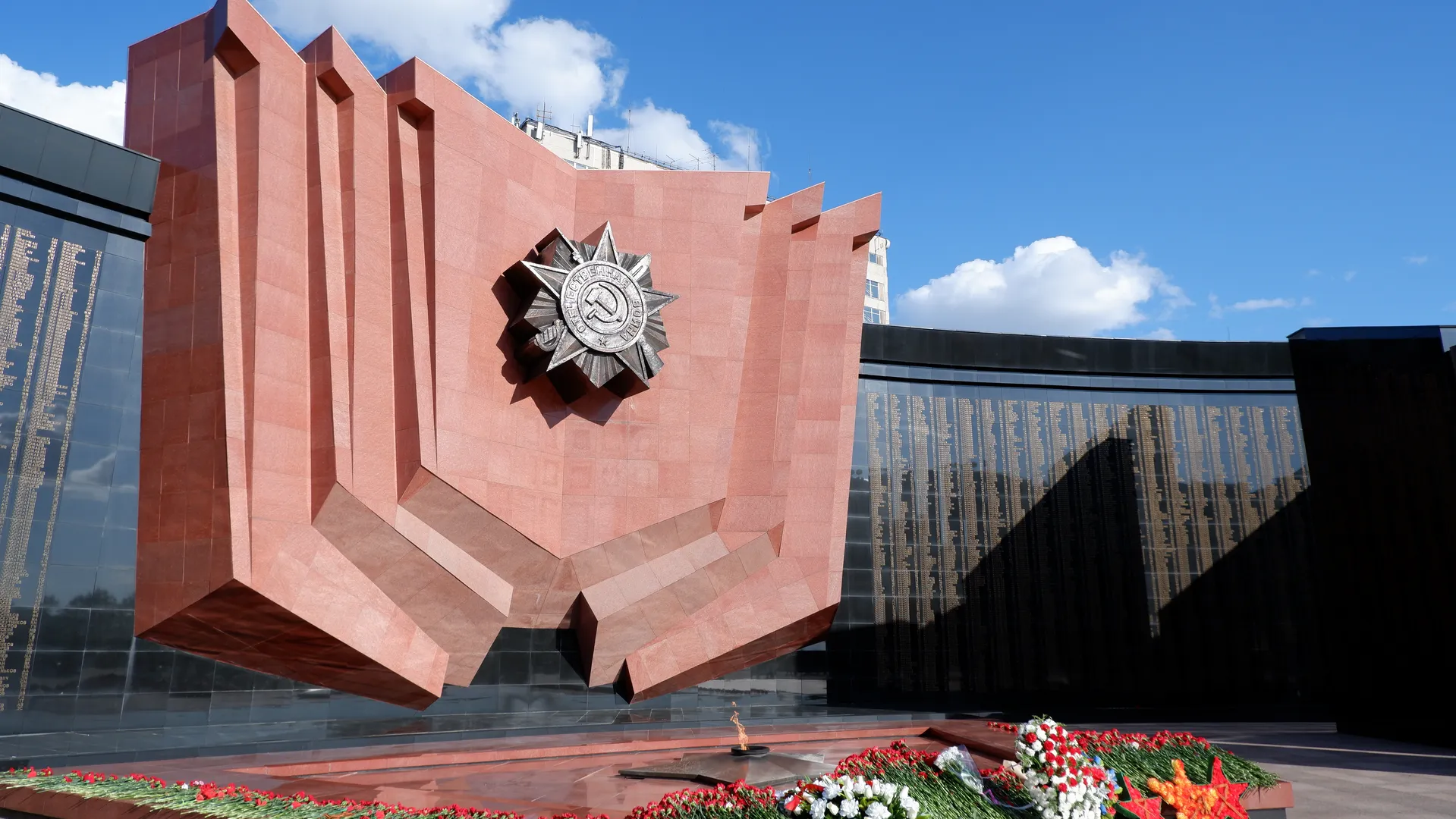 A red monument with a unique shape and a badge on top. On both sides of the monument are black stone slabs with engraved text. In front of the monument, there is a flower bed and a flame sculpture.