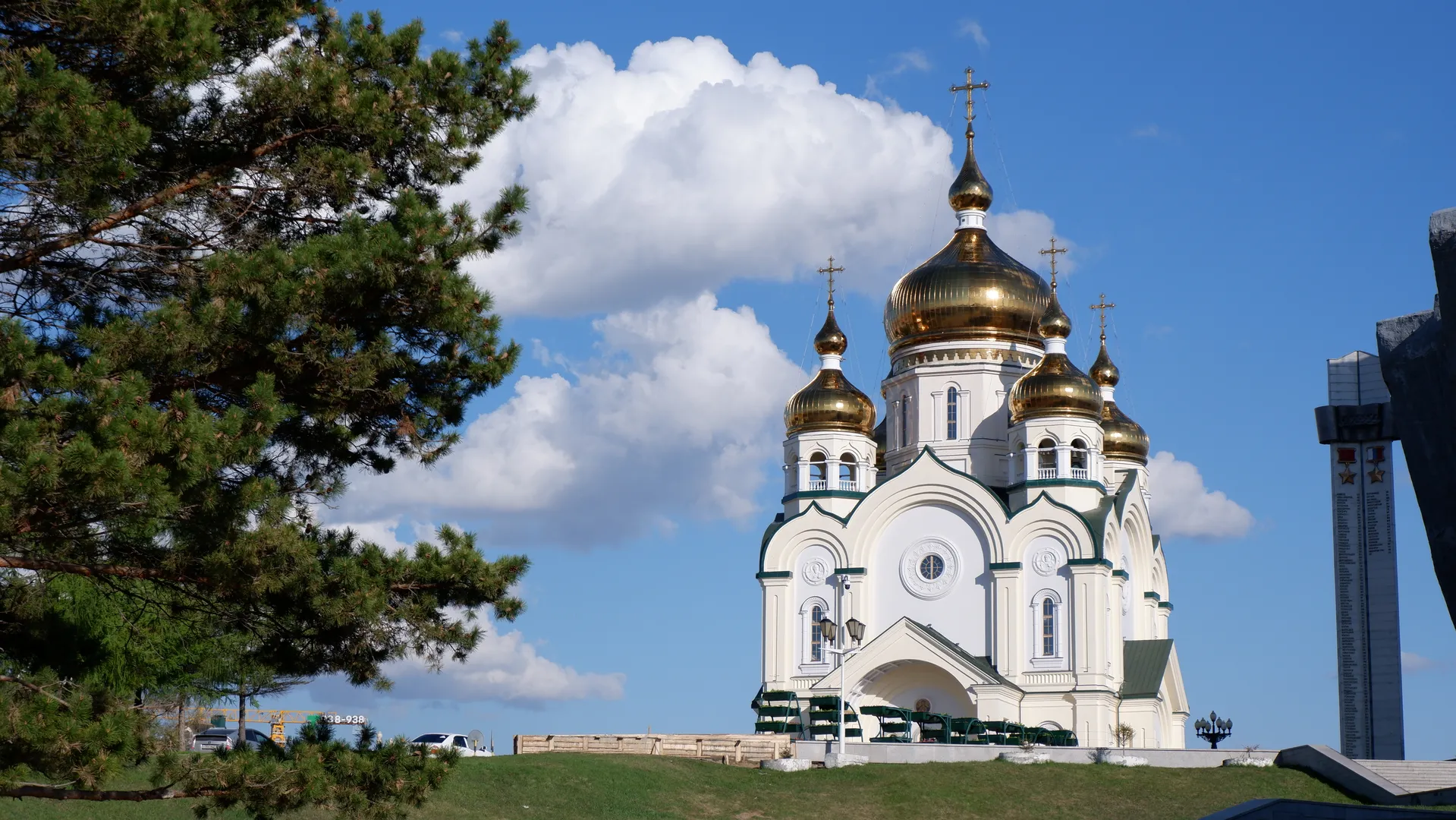 A white church with five golden domes and crosses on top. Dense pine trees are on the left side of the church, with a backdrop of blue skies and white clouds. In front of the church, there is a fence and vehicles.