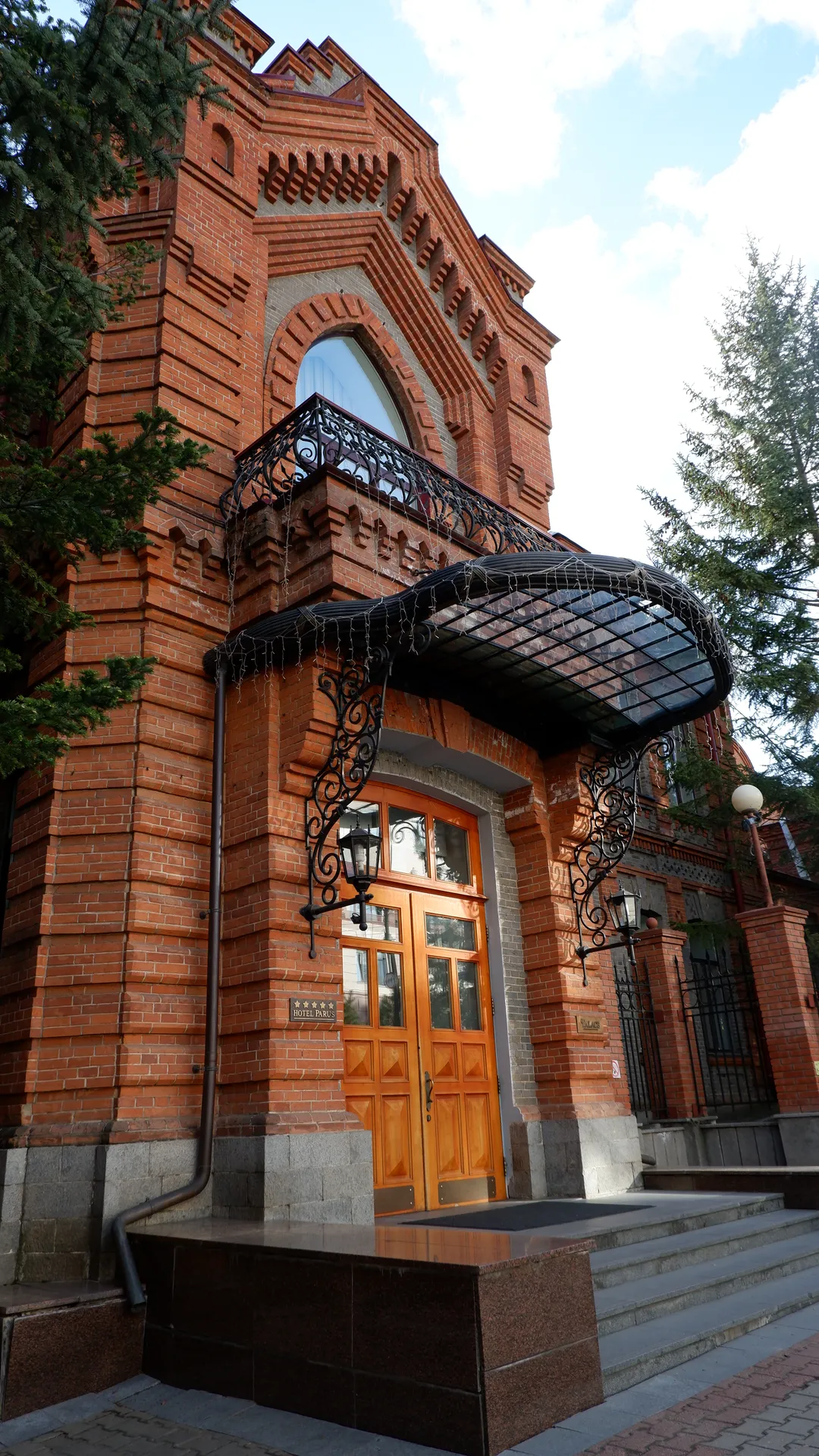 A red brick building with an orange wooden door and black wrought-iron decorations. The building’s front has arched windows and a balcony. Stairs and street lamps are in front of the building.