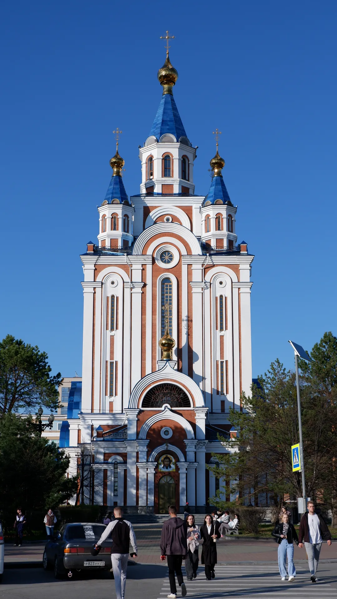 A church with a golden dome and a blue roof stands under the blue sky. The church’s front has multiple arched windows and decorative columns. People are walking in the square in front of the church.
