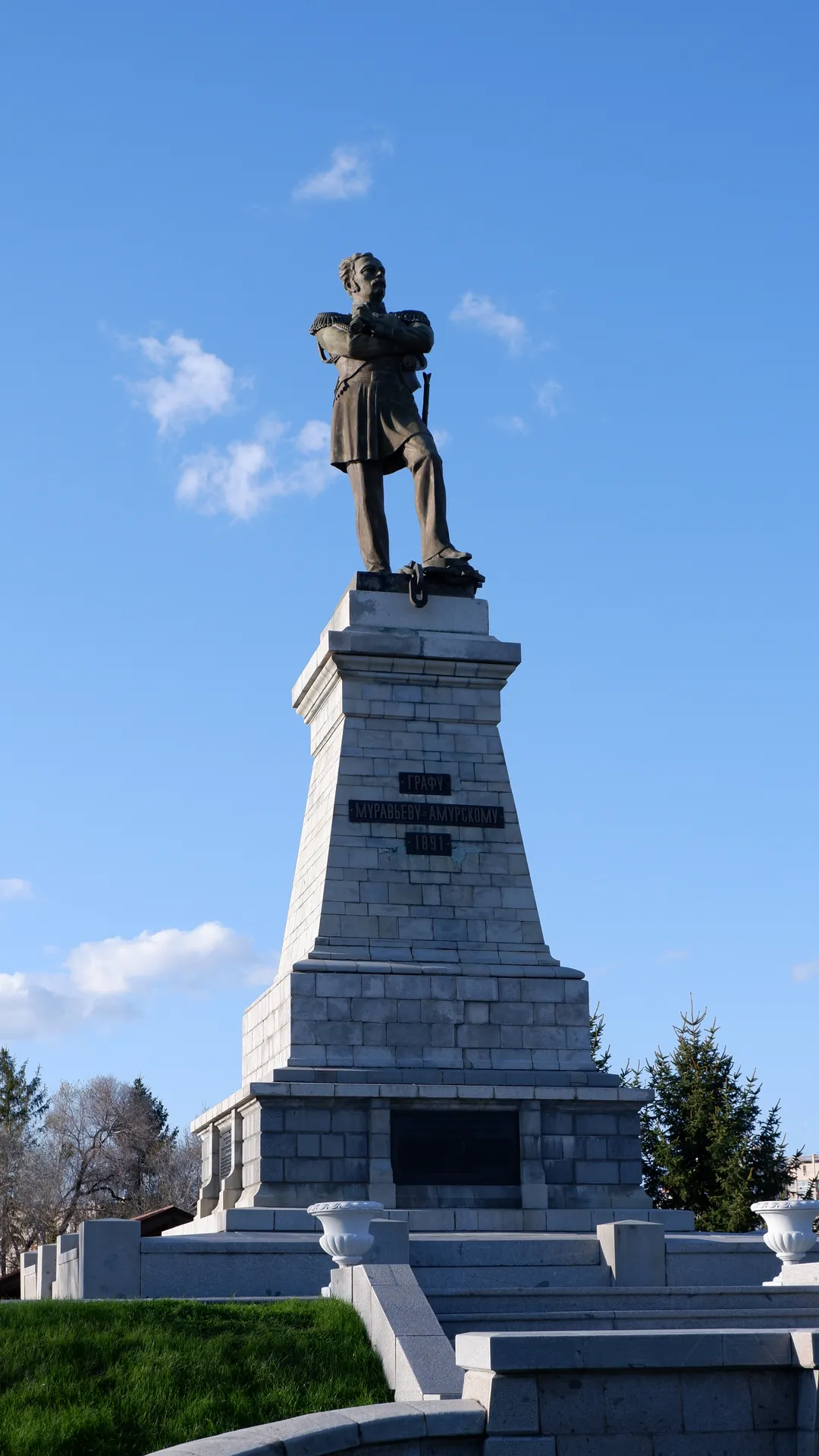 A statue stands on a stone pedestal, with the figure’s hands crossed. There is an inscription in Russian on the pedestal, and the background features a clear blue sky. A few trees surround the statue.