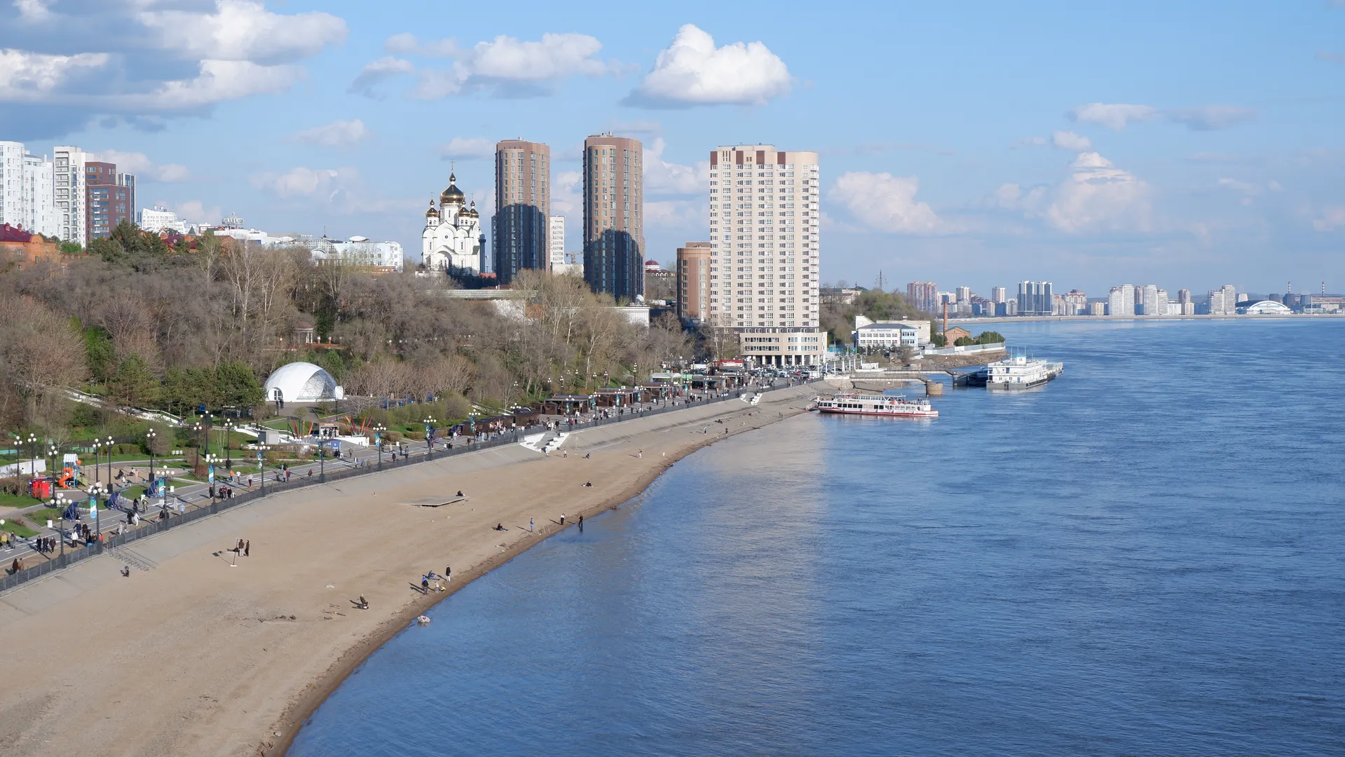 A river flows alongside a sandy beach, with pedestrians and facilities on the sand. In the distance, there are high-rise buildings and a golden-domed church. A few white clouds are in the sky.