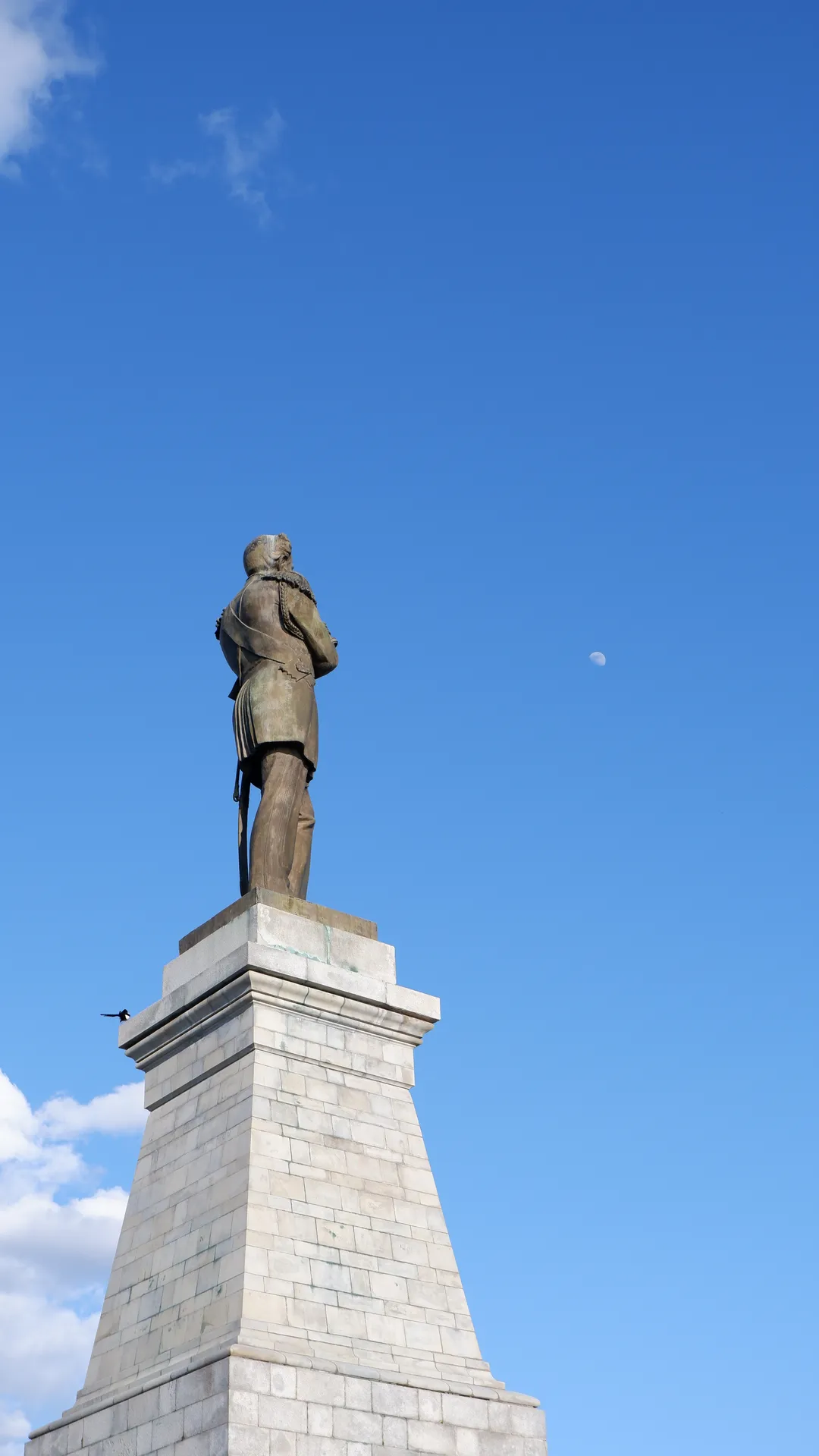 A statue stands on a tall stone pedestal, facing to one side. The background features a clear blue sky with a full moon in the air. The statue’s posture is upright.