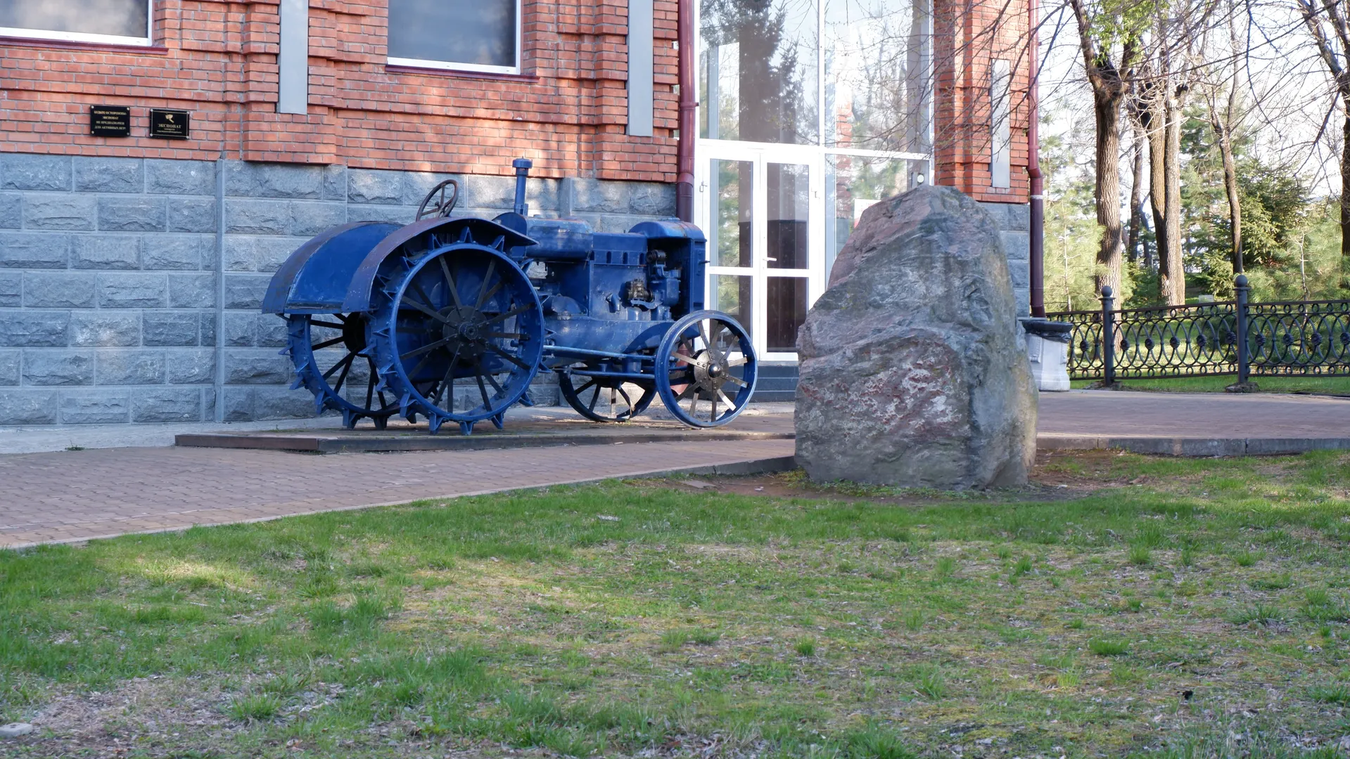 A blue vintage tractor is parked in front of a red brick building. Next to the tractor, there is a large stone, and the ground is covered with green grass. The glass door of the building reflects the light.