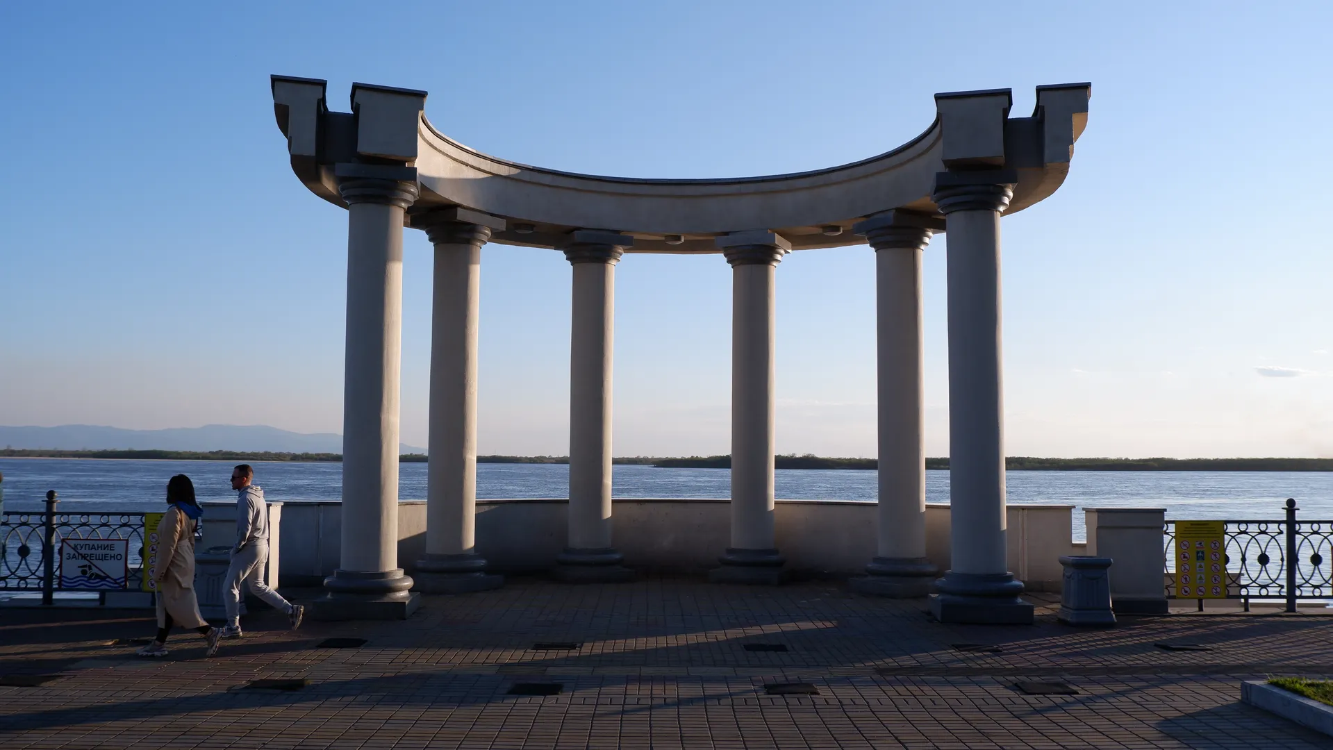 A semicircular colonnade stands by the water, supported by multiple white columns. Two people walk in front of the colonnade, with a vast expanse of water and distant mountains in the background. The sky is clear.