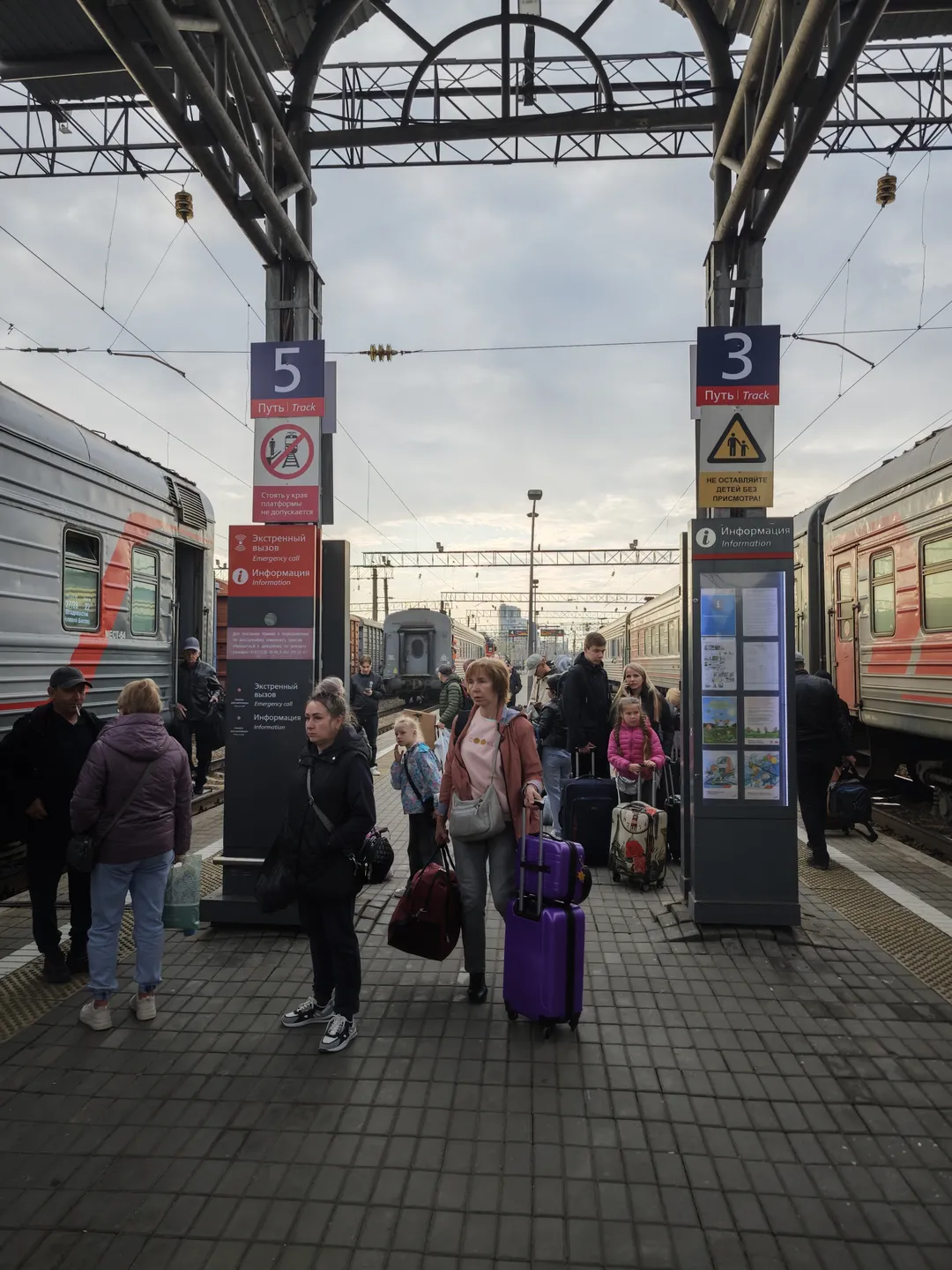 A crowded train platform with many people carrying suitcases. Two trains are parked on either side of the platform, with tracks and overhead power lines visible. There are signboards and an information display screen on the platform.