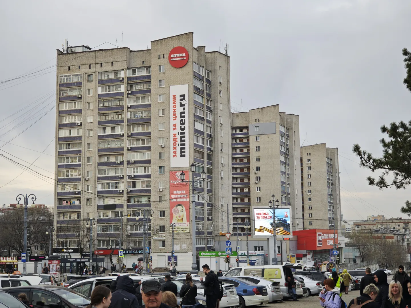 An old high-rise building with a gray exterior and neatly arranged windows. Multiple billboards are placed on the building. Vehicles are parked in the foreground, and many people are walking along the street in front of the building.
