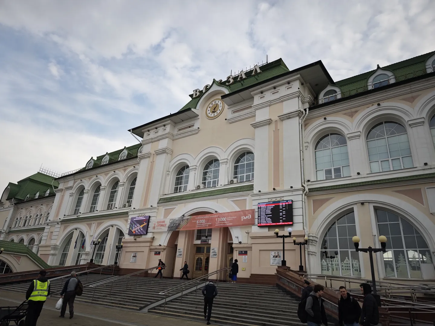 A large building with a green roof and a golden clock, featuring many arched windows on the front. There are stairs in front of the building, and people are walking up and down. The sky is cloudy, and more buildings are visible in the background.