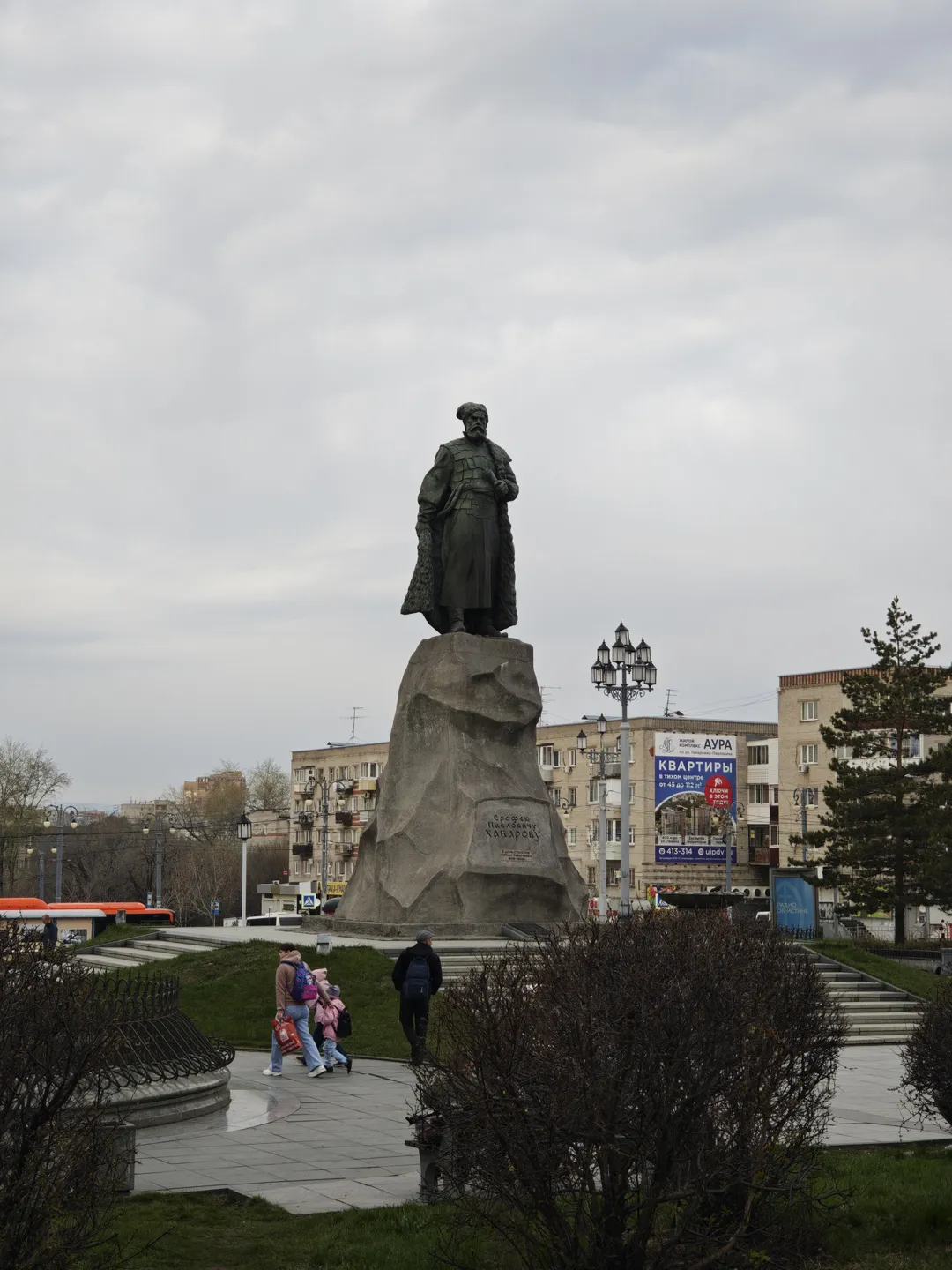 A statue stands on a stone pedestal, with the figure wearing a robe and holding an object. Shrubs and trees surround the statue, and multi-story buildings and street lights are visible in the background. The sky is overcast, and the ground is neatly paved.