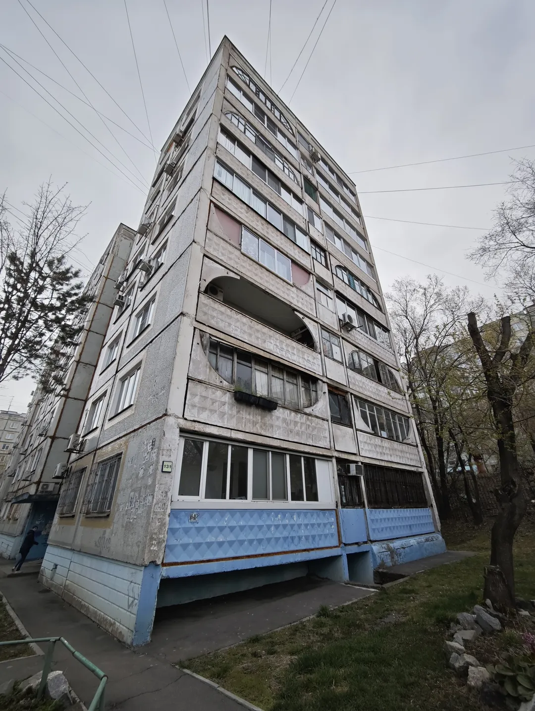 A high-rise building with a light-colored exterior and blue decorations at the bottom. The building has multiple balconies and windows, with slight stains on the exterior. Trees and power lines surround it, and there is a path and grassy area on the ground.