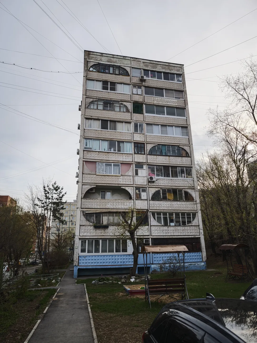 A high-rise building with a light-colored exterior, featuring arched balconies on each floor. The building has blue decorations at the base, with trees and greenery around it. A small path leads in front, with a car parked nearby.