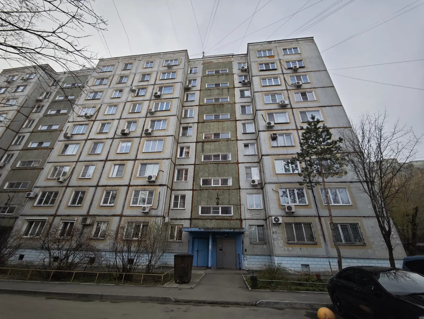 A multi-story building with a light gray exterior and neatly arranged windows. The entrance features a blue door, and there are trash bins and trees in front. A black car is parked nearby, and the ground is paved with concrete.