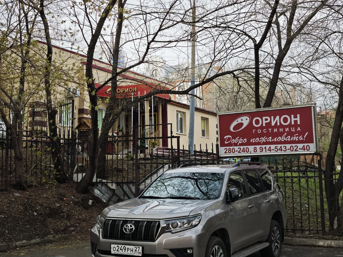 A silver SUV is parked in front of an iron fence, behind which is a building. The building has a red sign above it with the text “ОРИОН.” Trees surround the area, and the ground is covered with fallen leaves.