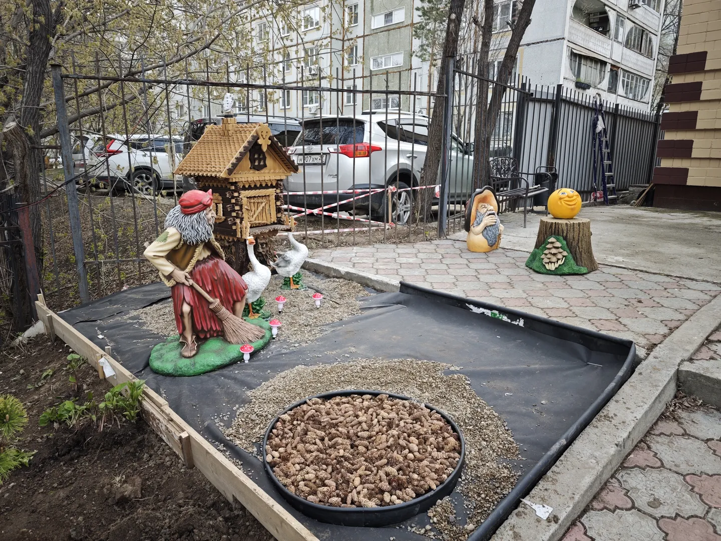 A decorative area with a black pot filled with pine cones at the center. Nearby is a sculpture of a person holding a broom and two geese. In the background, parked vehicles and high-rise buildings are visible.