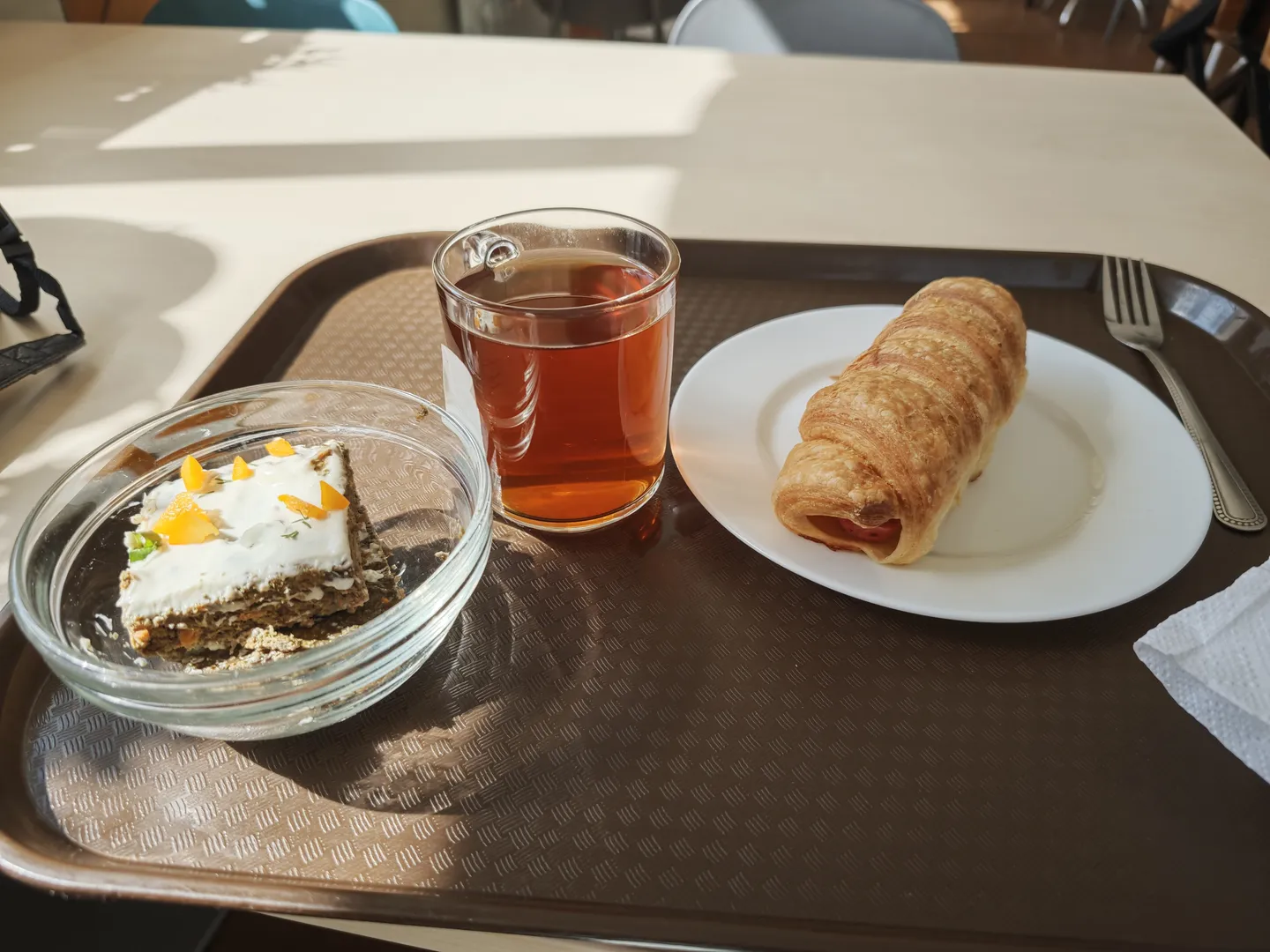 A tray with a cup of tea, a croissant, and a piece of cake. The transparent tea cup holds the drink, the croissant is golden brown, and the cake is decorated with cream and orange slices. The tray is brown and placed on a white table.