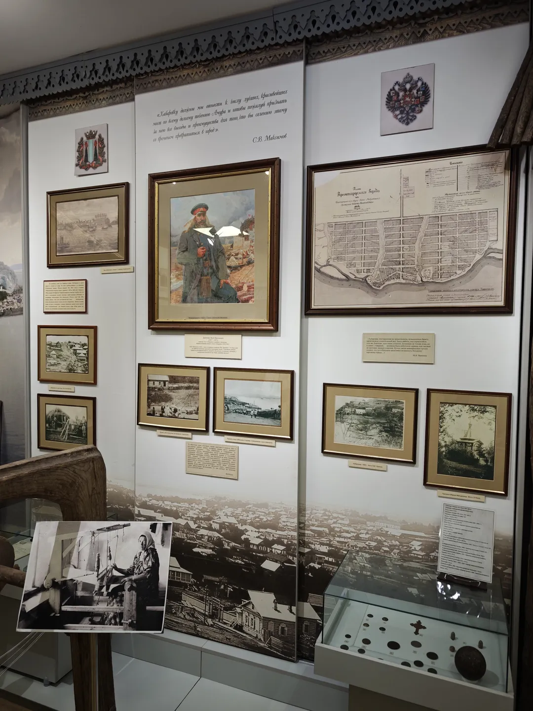 The wall is adorned with several paintings and photographs, with a display case below. The artworks and photos showcase historical scenes and maps. The display case contains small exhibits, and there are explanatory texts on the wall.