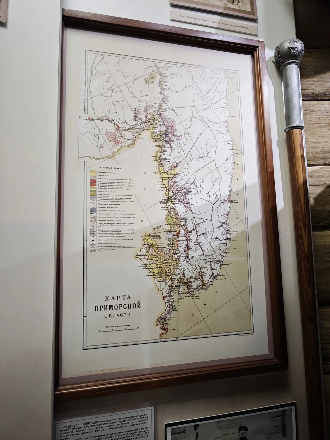 A framed map hangs on the wall, with various color-coded markings. Below the map is a Russian title, and next to it stands a decorative pillar. Other exhibits and explanatory texts are visible on the wall.