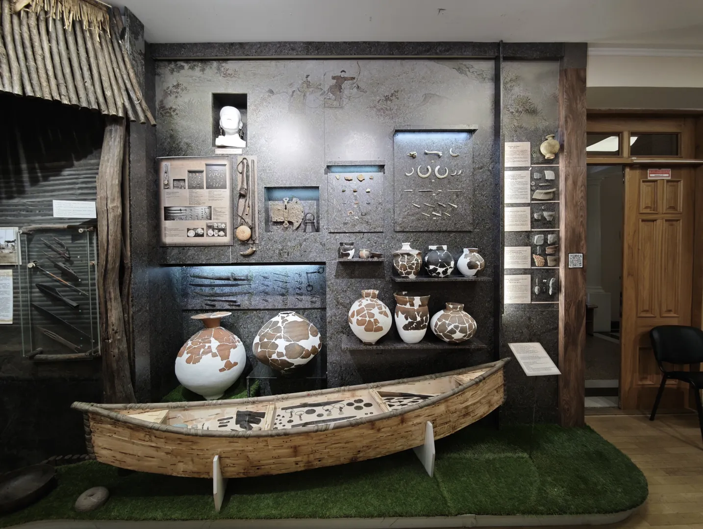 The museum display area features ancient pottery and tools, with informational plaques hanging on the walls. A wooden canoe is placed on a green carpet, with a large ceramic pot next to it. The background wall displays murals and sculptures.