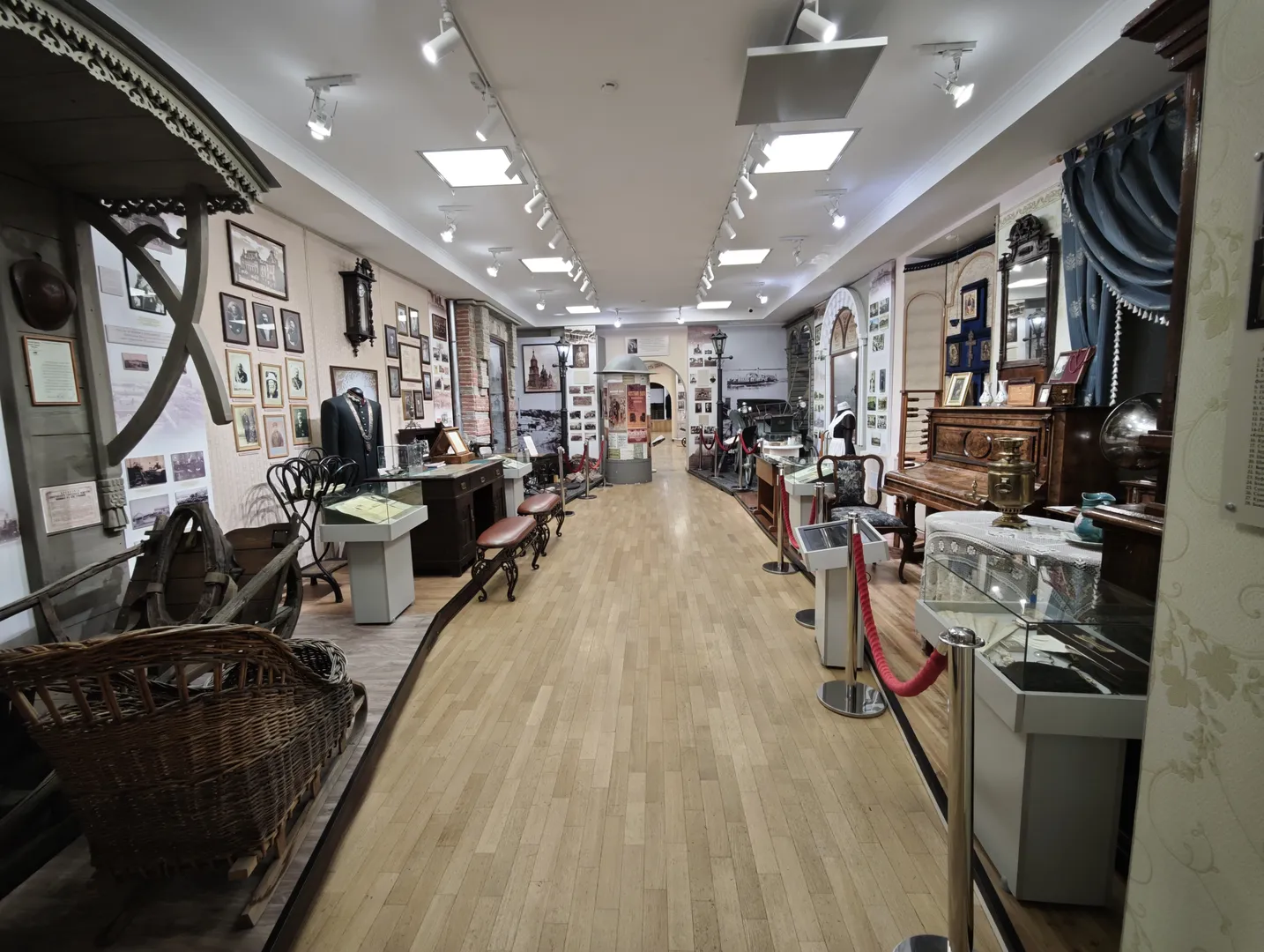 The museum corridor is lined with historical artifacts and furniture, with wooden flooring. The walls are filled with photographs and framed artworks, and display cases contain various items. The central passage is wide and well-lit, guiding visitors forward.