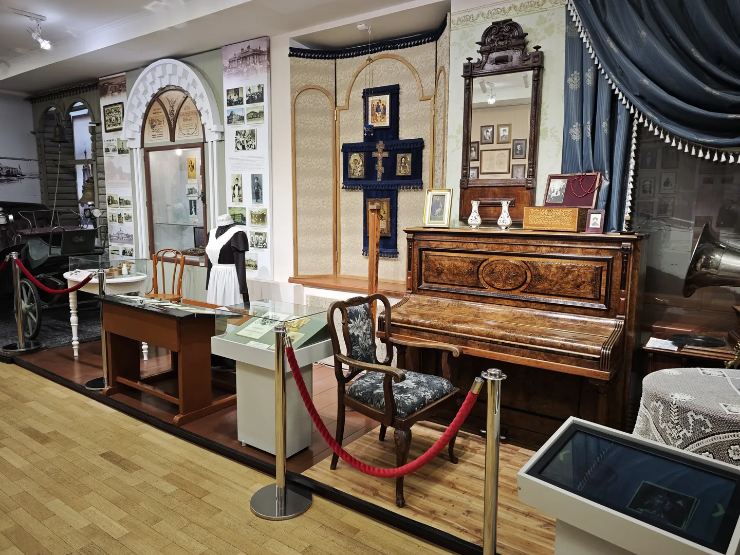 In the room, an antique piano and matching chair are displayed, with a mirror and decorations hanging above the piano. A red rope barrier protects the exhibit from being touched. The background wall is adorned with religious portraits and picture frames.