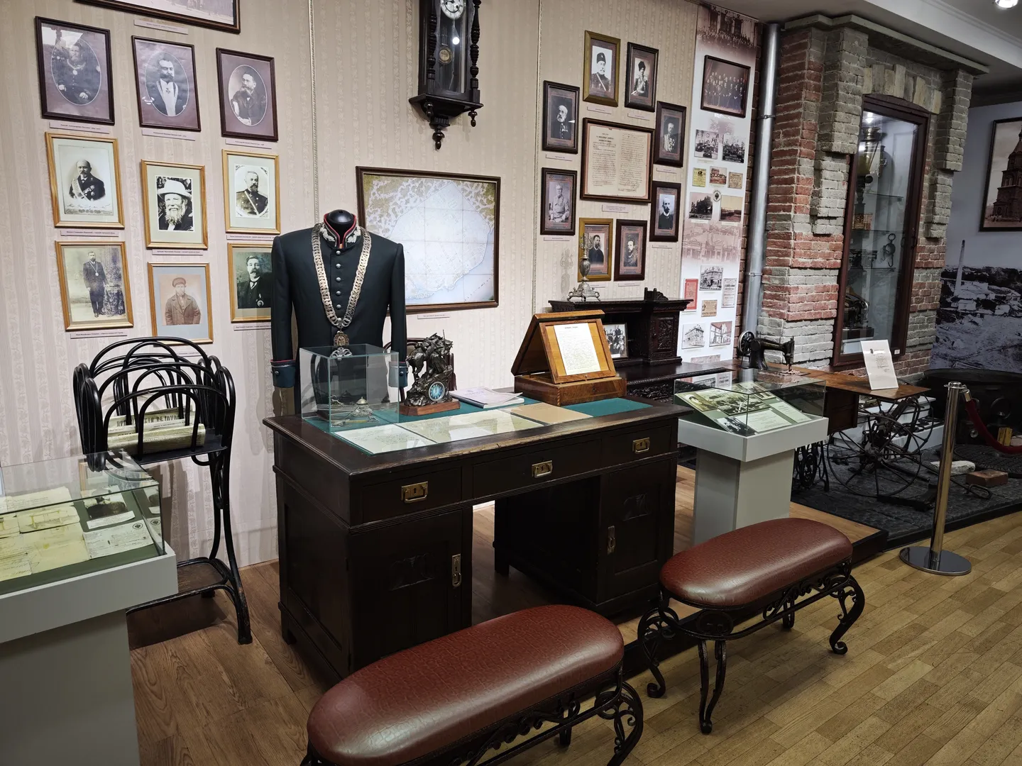 The exhibition area features a vintage desk with documents and seals on it. Behind the desk stands a mannequin dressed in a uniform, with historical photographs hanging on the wall. A nearby display case contains a sewing machine and other items.