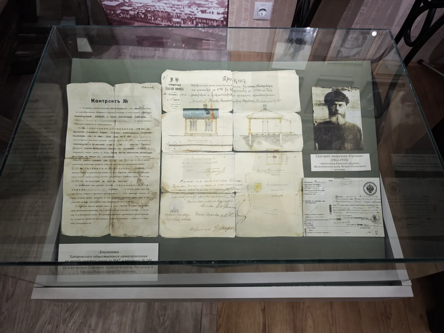 A glass display case showcases several ancient documents and a photograph of a person, with handwritten text and seals on the documents. The base of the display case has a label explaining the historical context of the documents. The background wall features an image of a building.