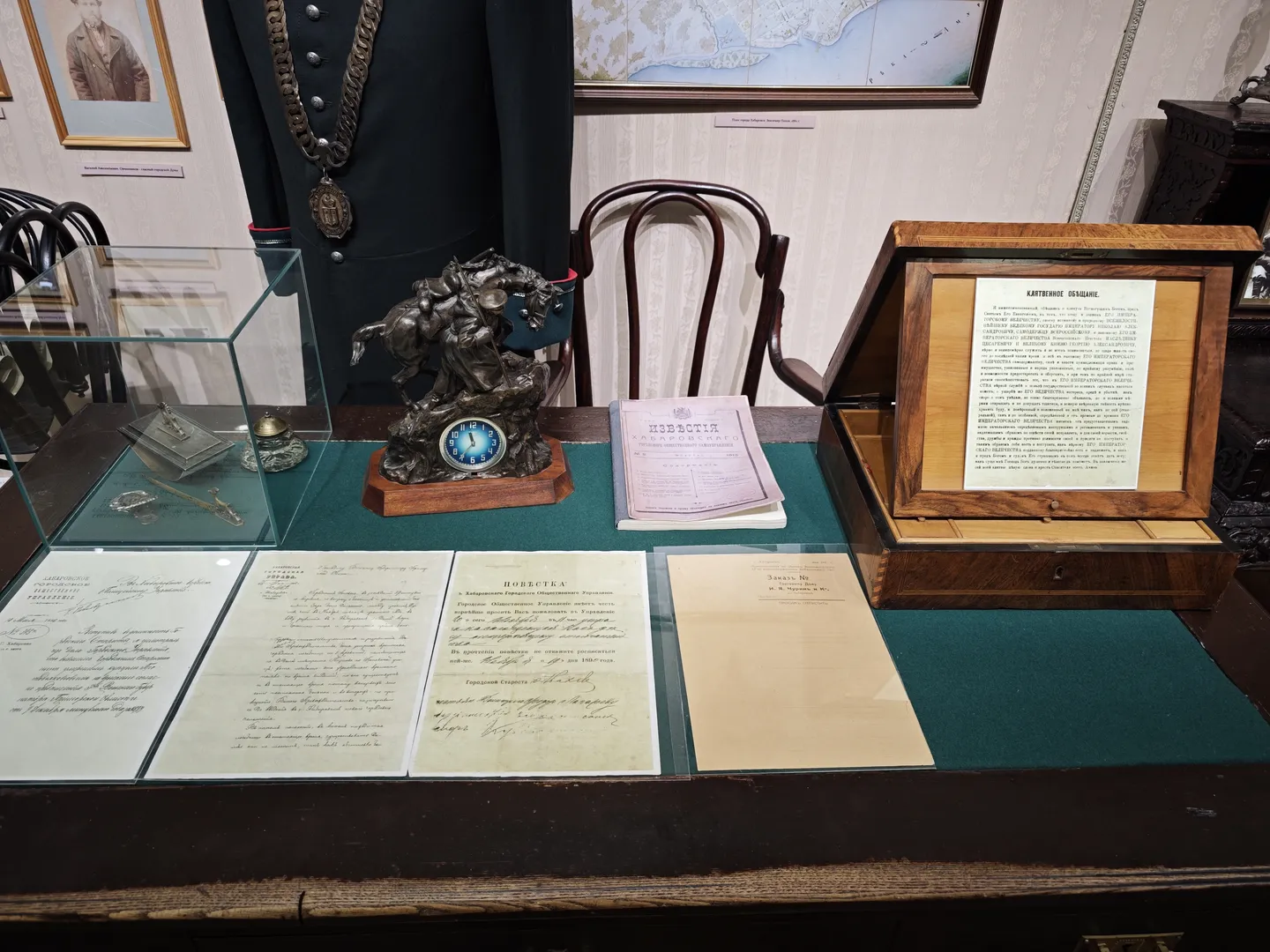 On the table are several documents and a decorative clock sculpture. The documents are neatly arranged, covering historical records. The background wall features a map and a portrait painting of a person. A chair is placed in front of the table.
