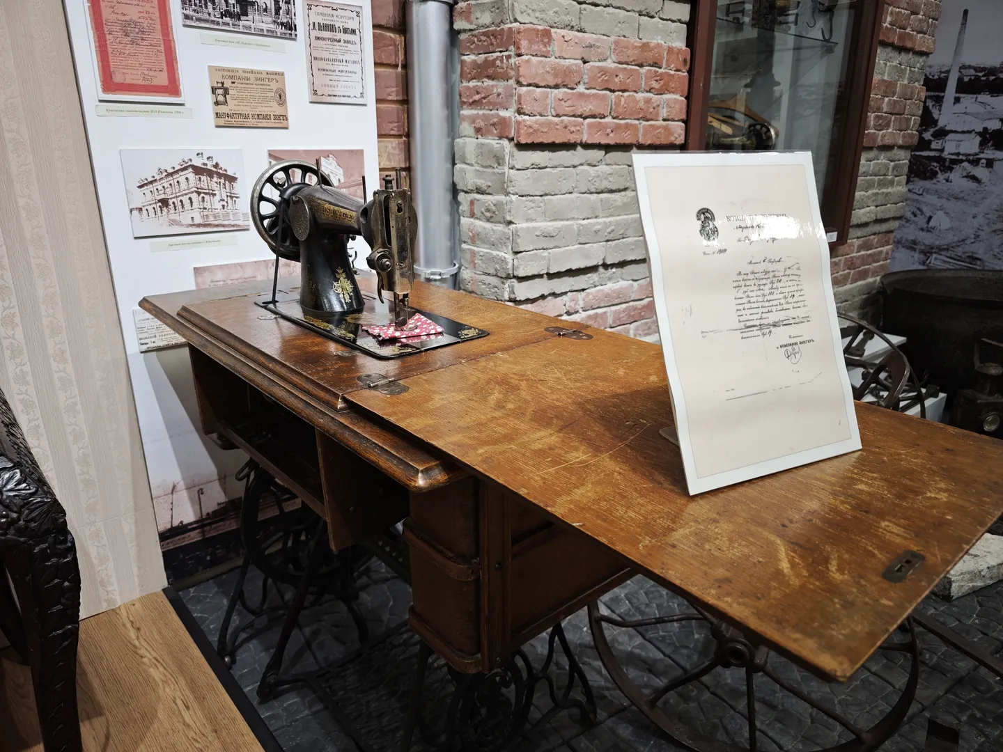 An antique sewing machine sits on a wooden table, with a document placed next to it. The background wall is made of brick, with historical photographs and posters affixed to it. The sewing machine design is intricate, showcasing early industrial craftsmanship.