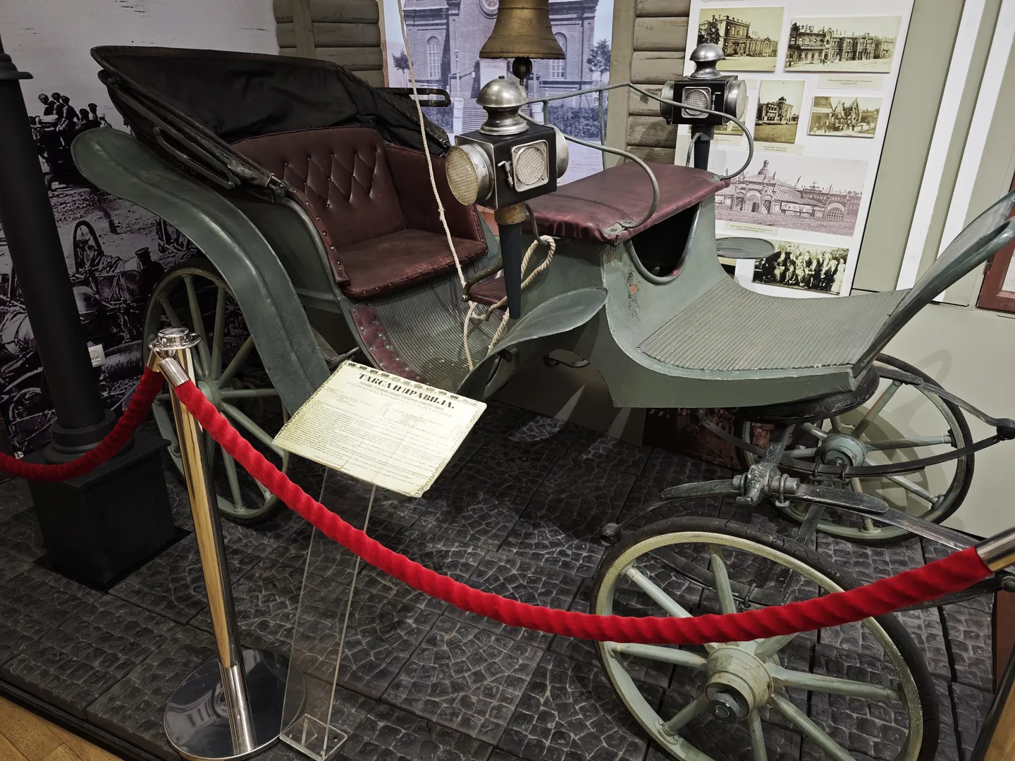 A vintage carriage is displayed in the museum, with its structure intact, complete with seats and a canopy. The front of the carriage has bells and lamps, with finely detailed elements. It is surrounded by a red rope barrier to protect the exhibit.